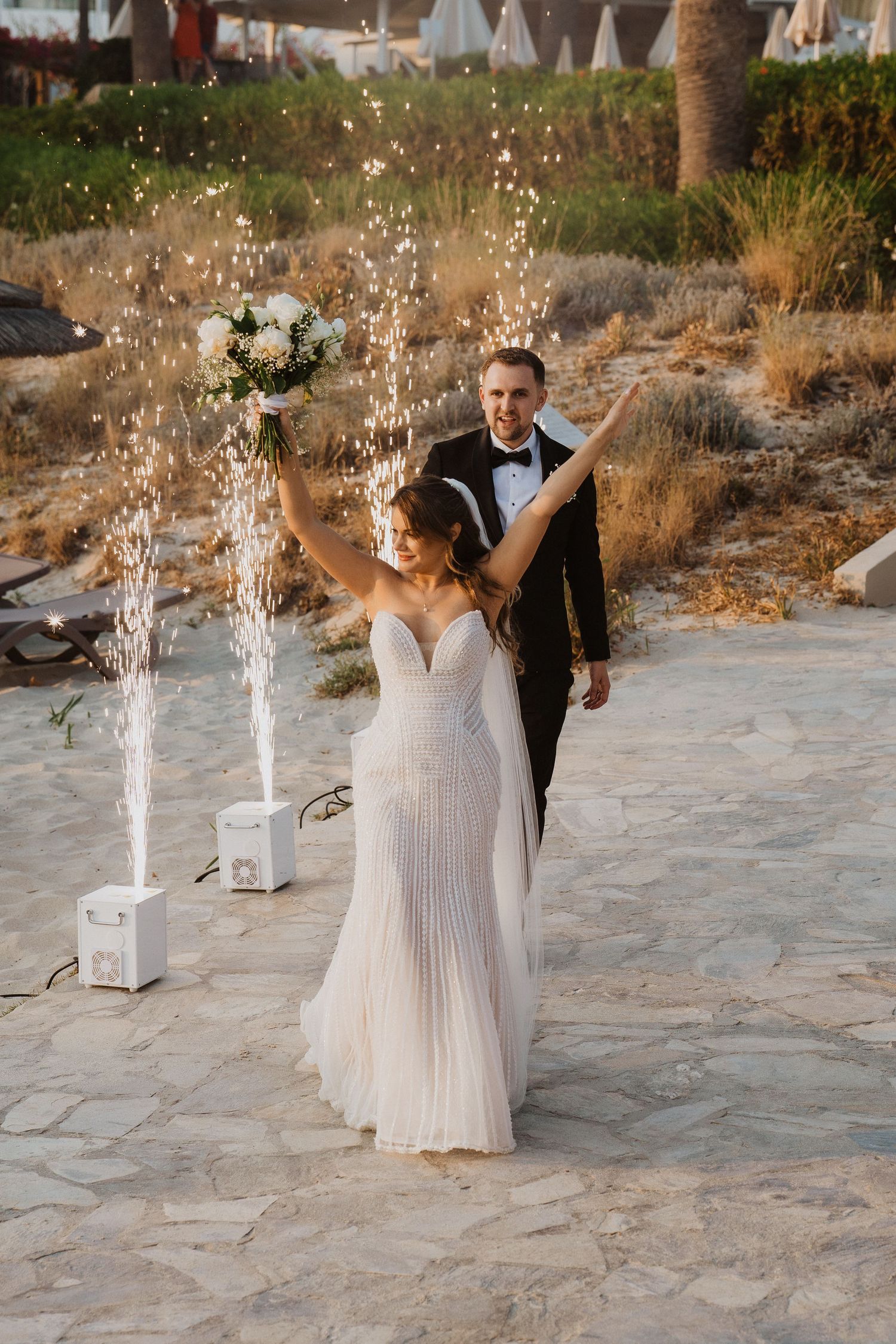 Bride raises bouquet joyfully as couple enters wedding reception amid sparkling fountain fireworks outdoors.