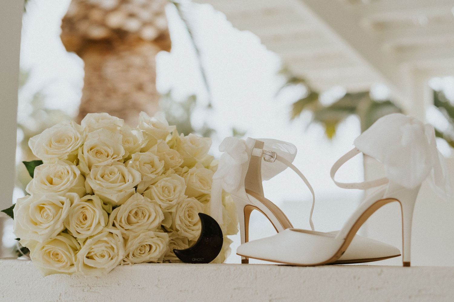 Elegant white wedding shoes next to cream rose bouquet on light surface.