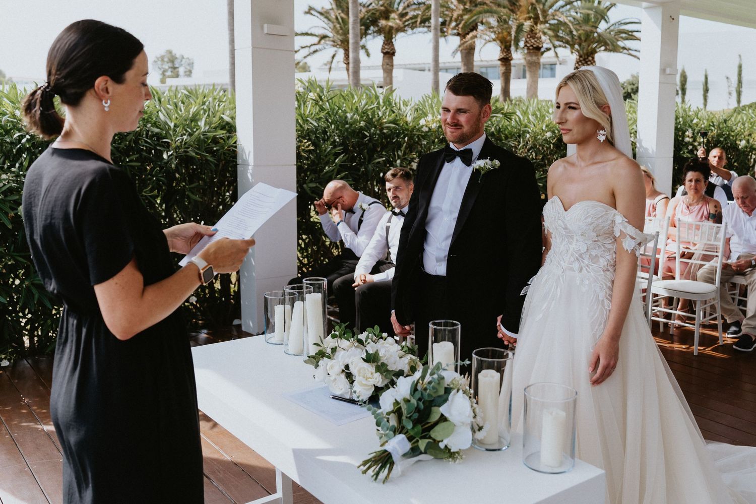 Wedding ceremony taking place on a covered porch with palm trees visible in the background and white floral arrangements.