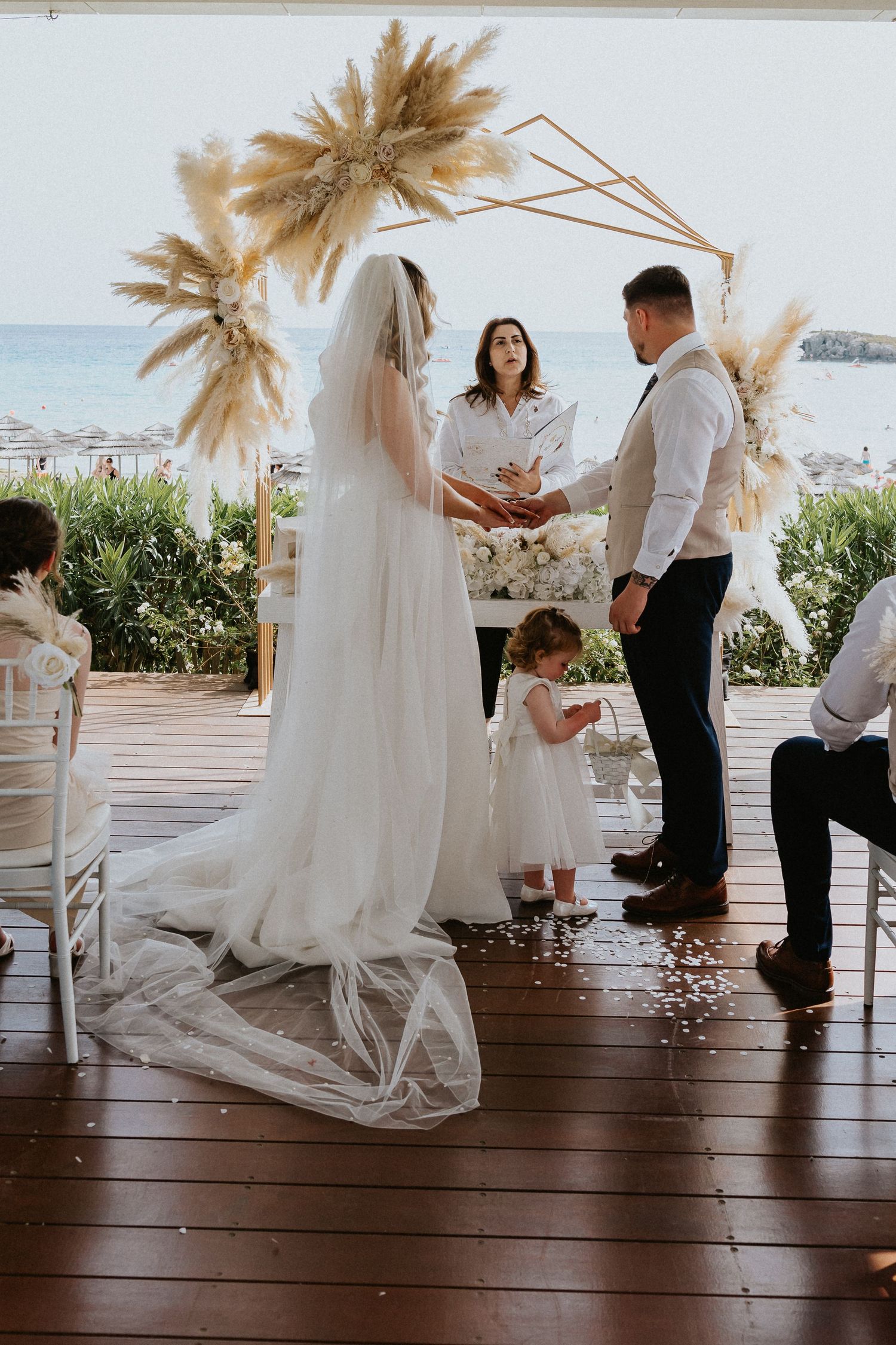 A beachside wedding ceremony under pampas grass decor with white florals and an ocean view backdrop.