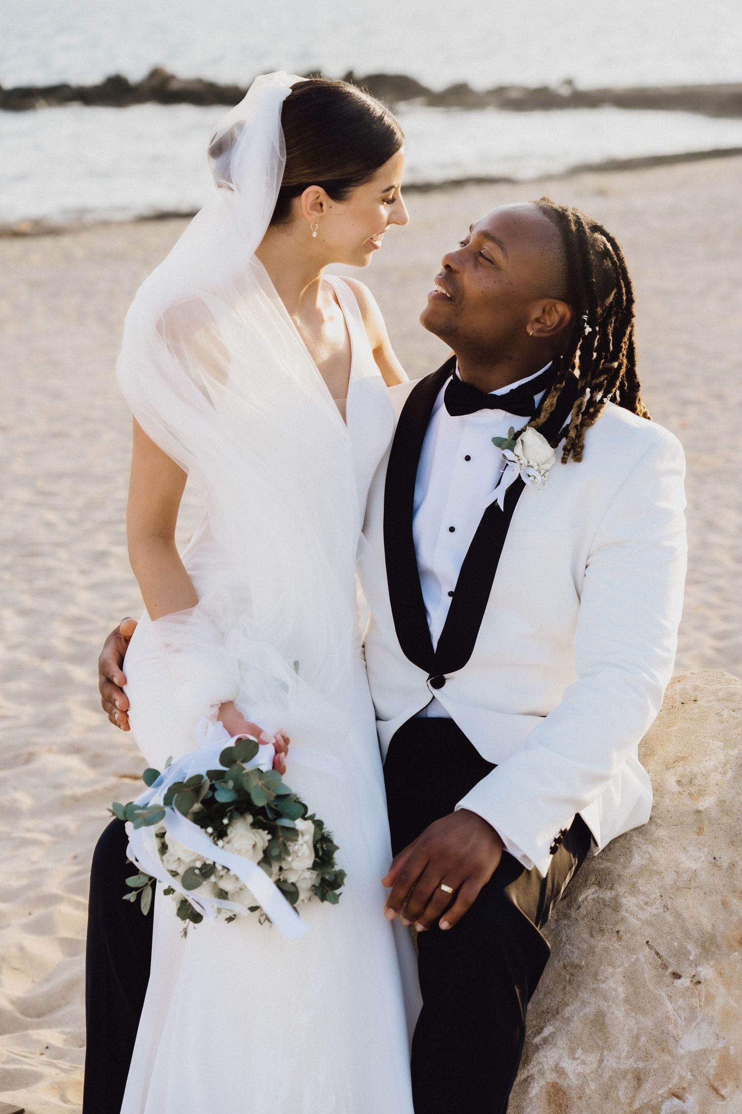 Wedding couple shares loving glance while holding bouquet against beach backdrop.