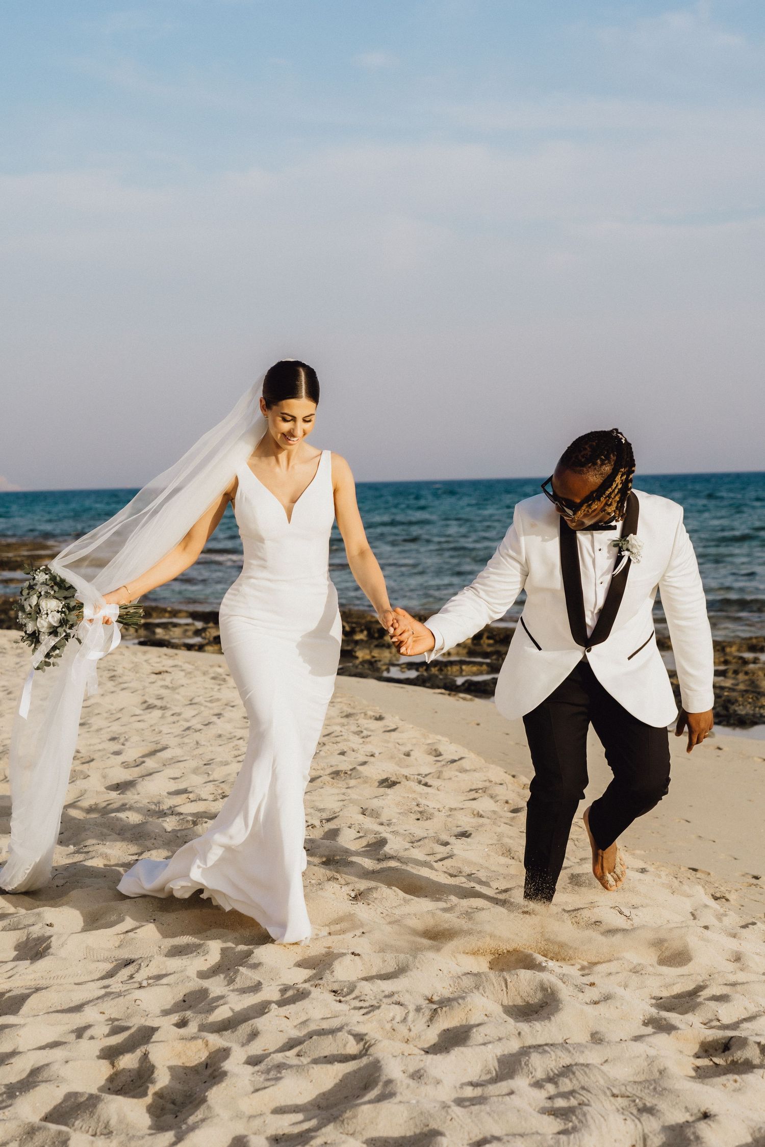 Couple walks hand in hand along beach in elegant wedding attire.