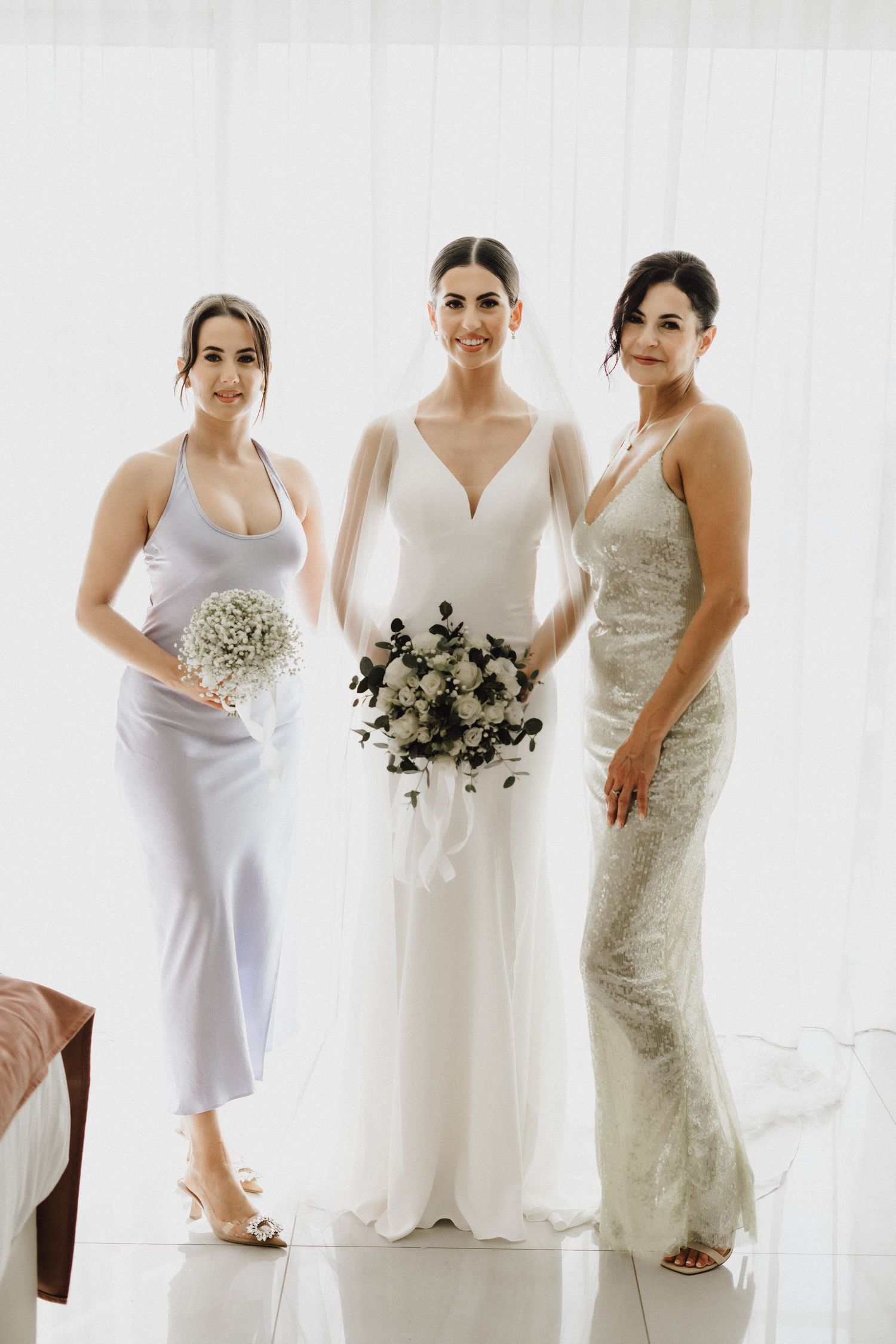 Three women in formal dresses pose together with wedding bouquets against a white backdrop.