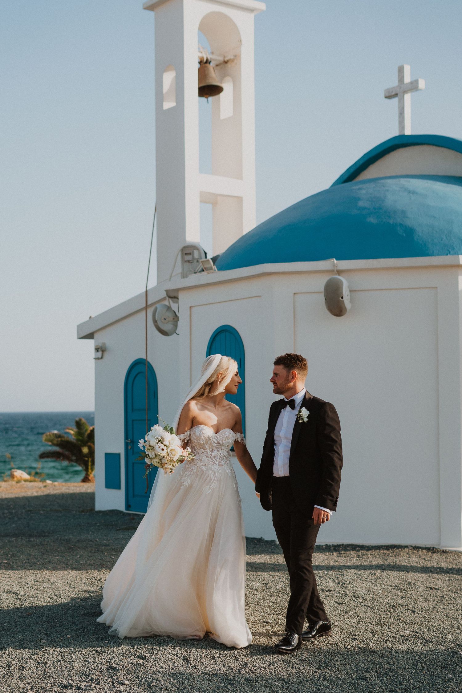 Bride and groom pose by traditional Greek chapel with blue dome and bell tower.