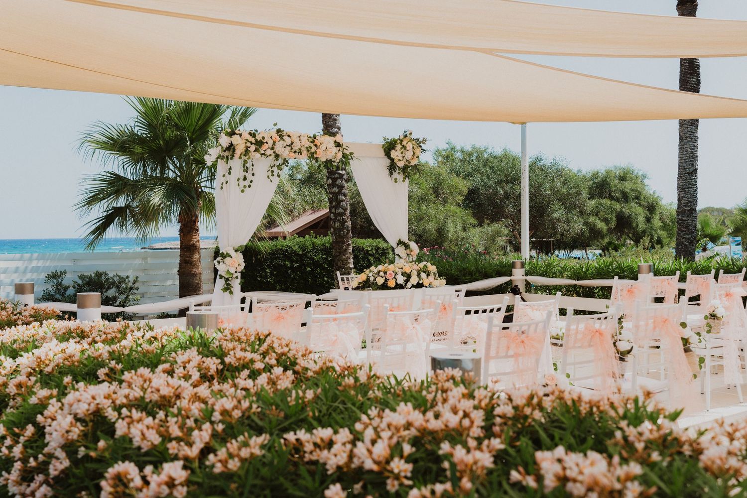 Outdoor wedding ceremony setup with sheer white curtains, flowers and chairs overlooking the ocean and palm trees.