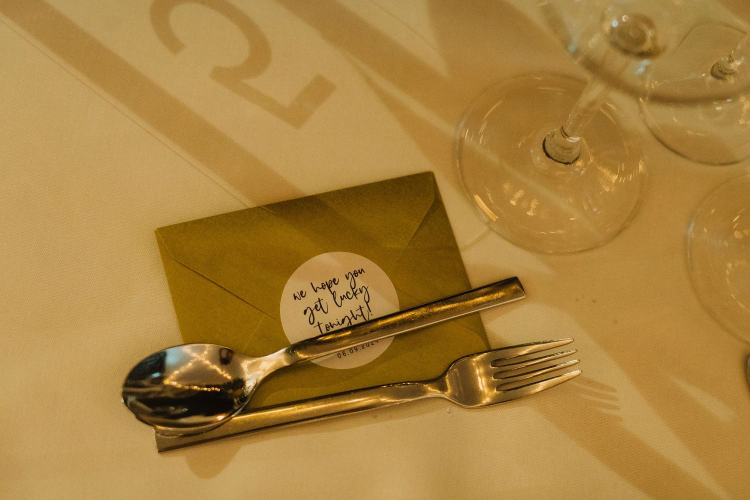 Close up of elegant place setting with gold envelope, cutlery and wine glasses on white tablecloth.