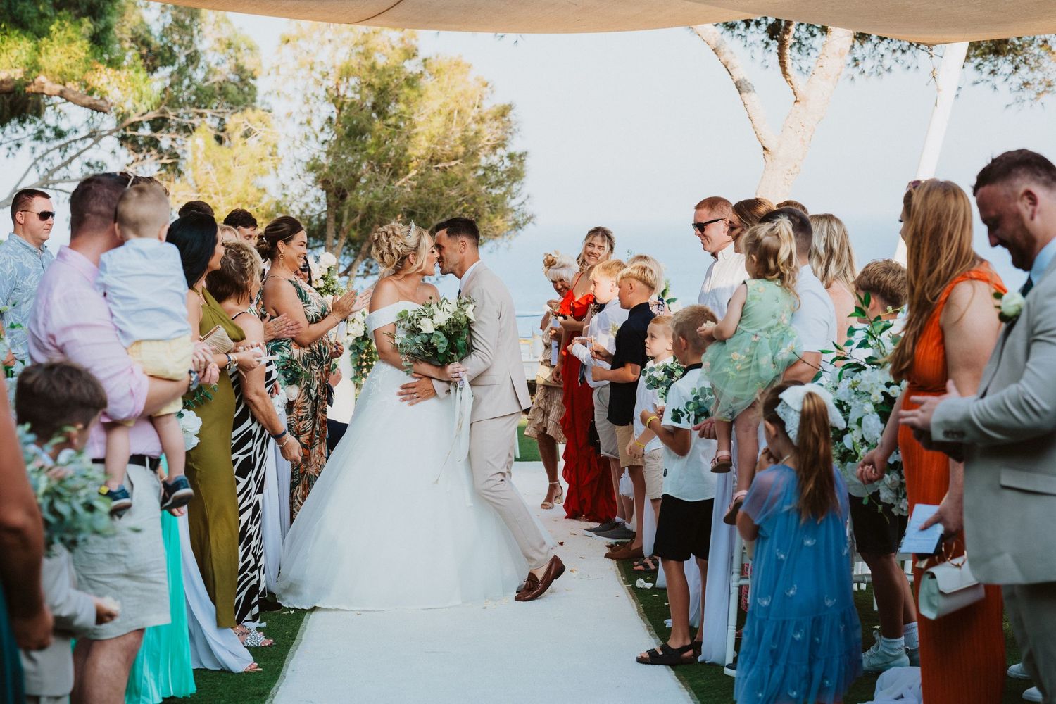 Wedding ceremony guests line a white aisle runner under tented canopy with greenery accents.