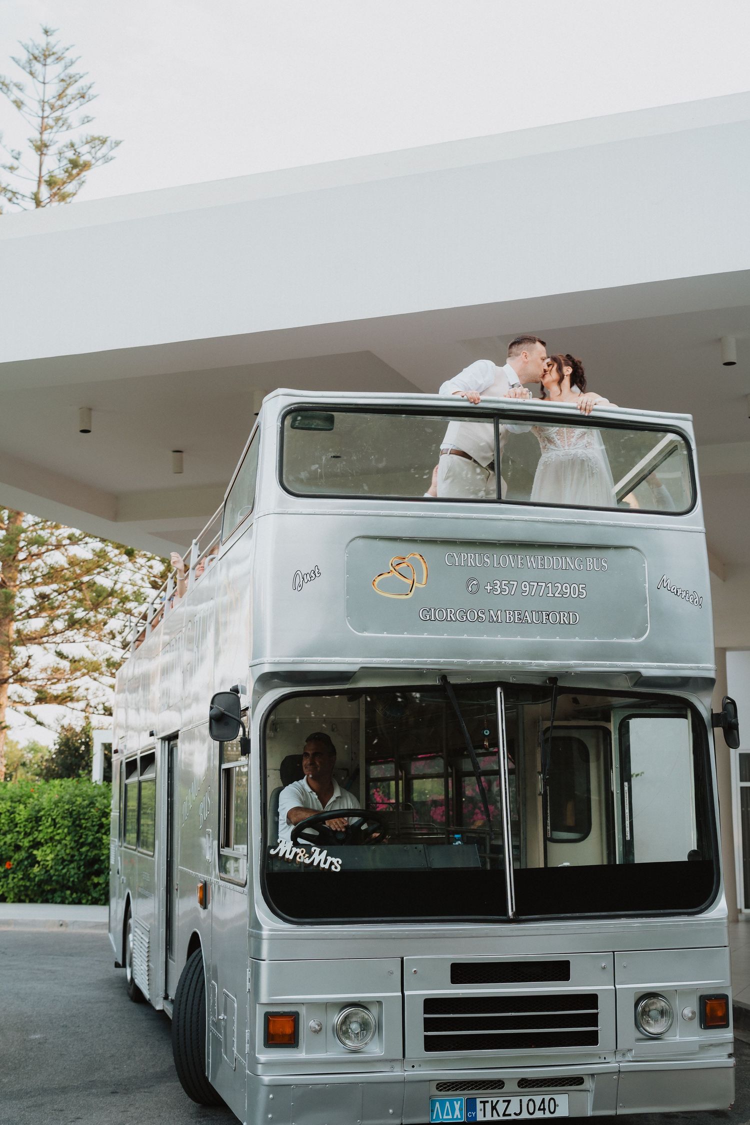 Vintage double-decker wedding party bus painted in light gray with a couple sharing a romantic moment on the top deck.