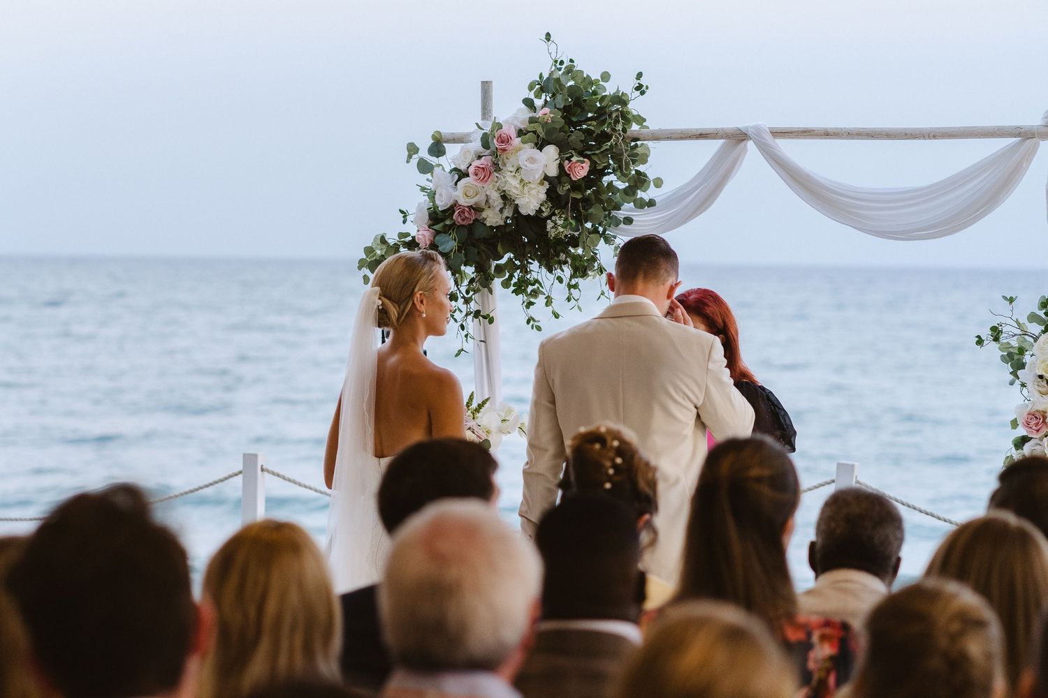Wedding ceremony by the ocean with floral arch and guests seated in covered venue.