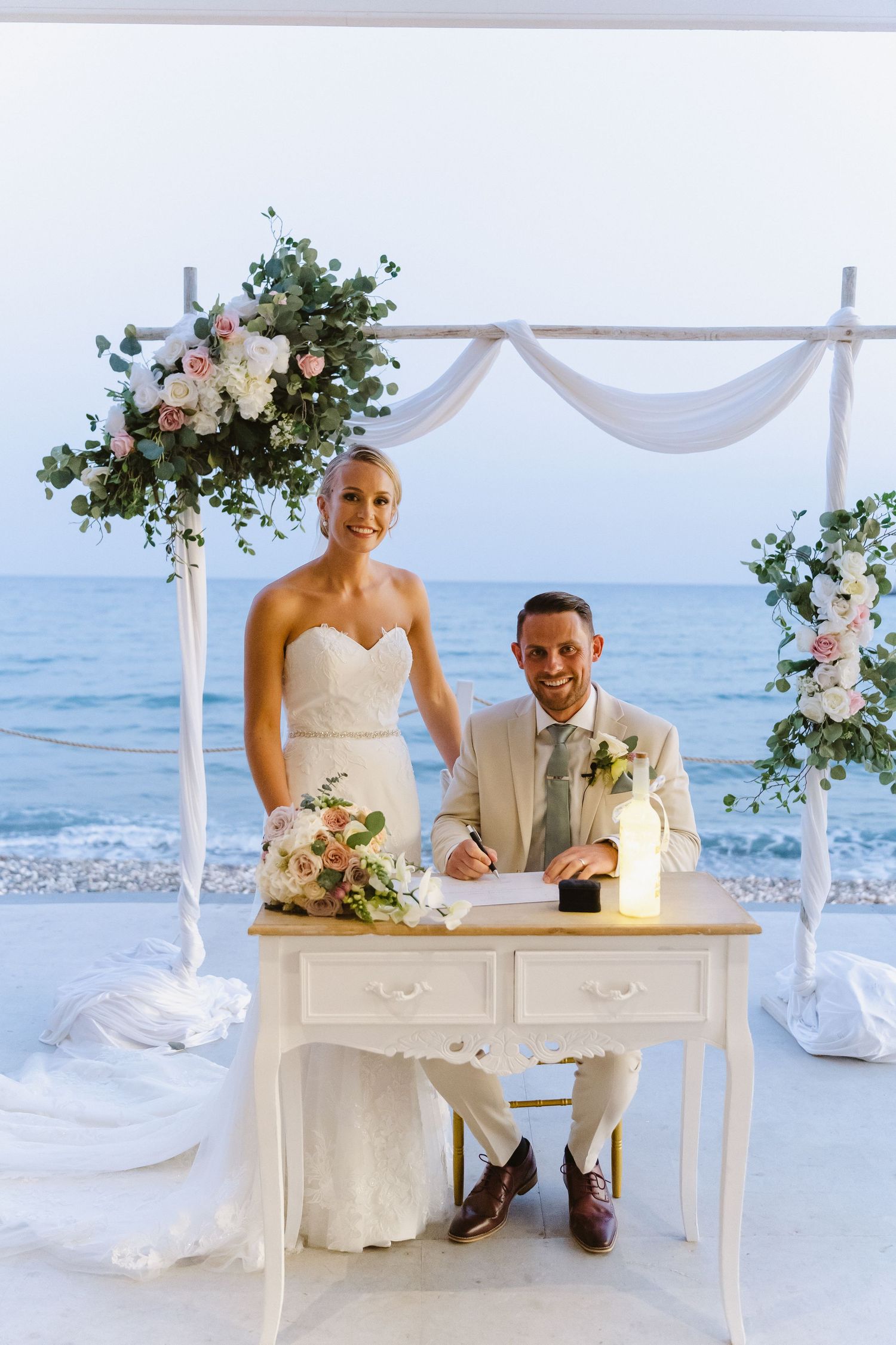 A beach wedding ceremony setup with white arch, floral arrangements, and ocean backdrop.
