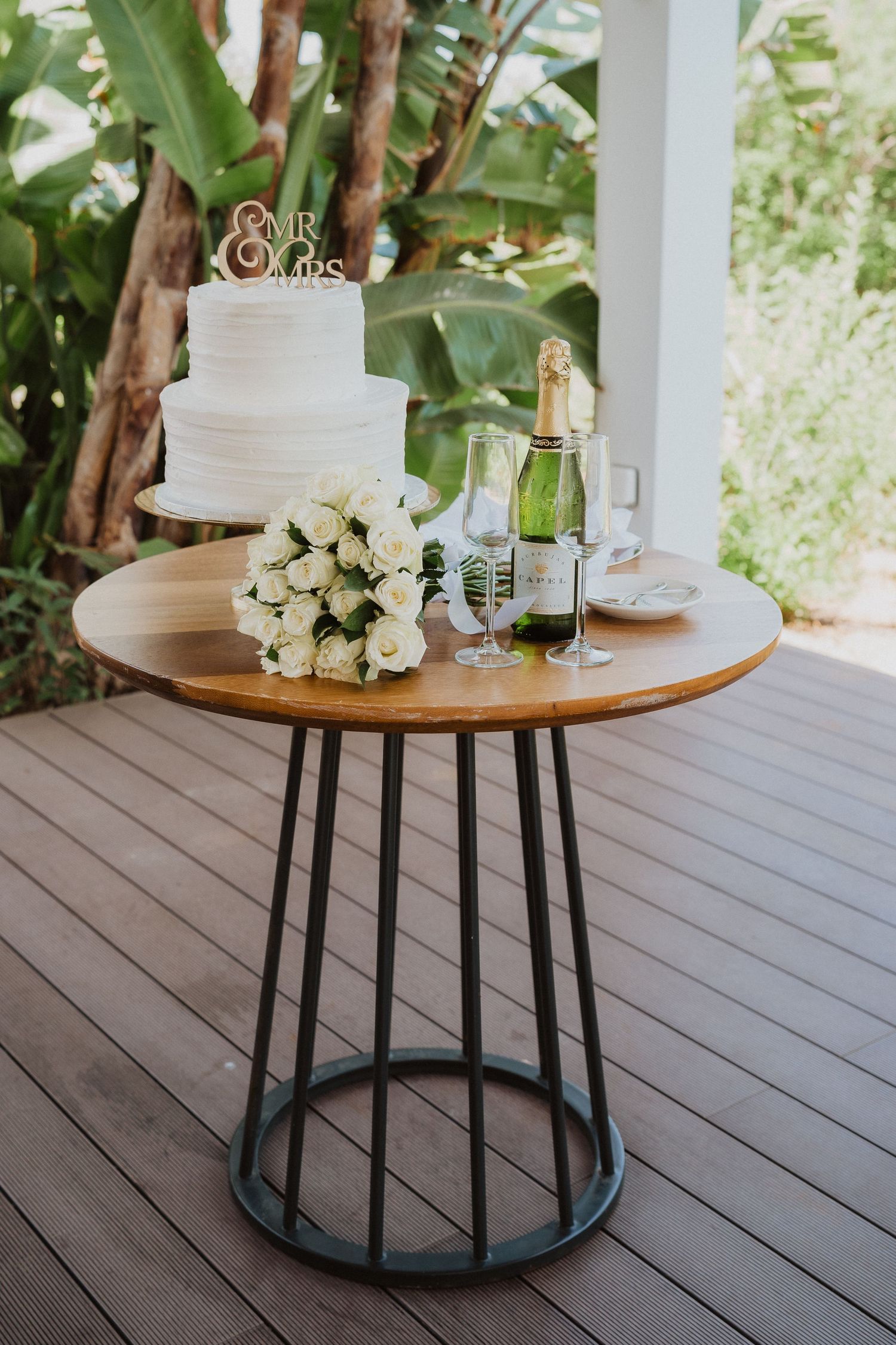 White wedding cake and bouquet displayed on round wooden table with champagne bottle.