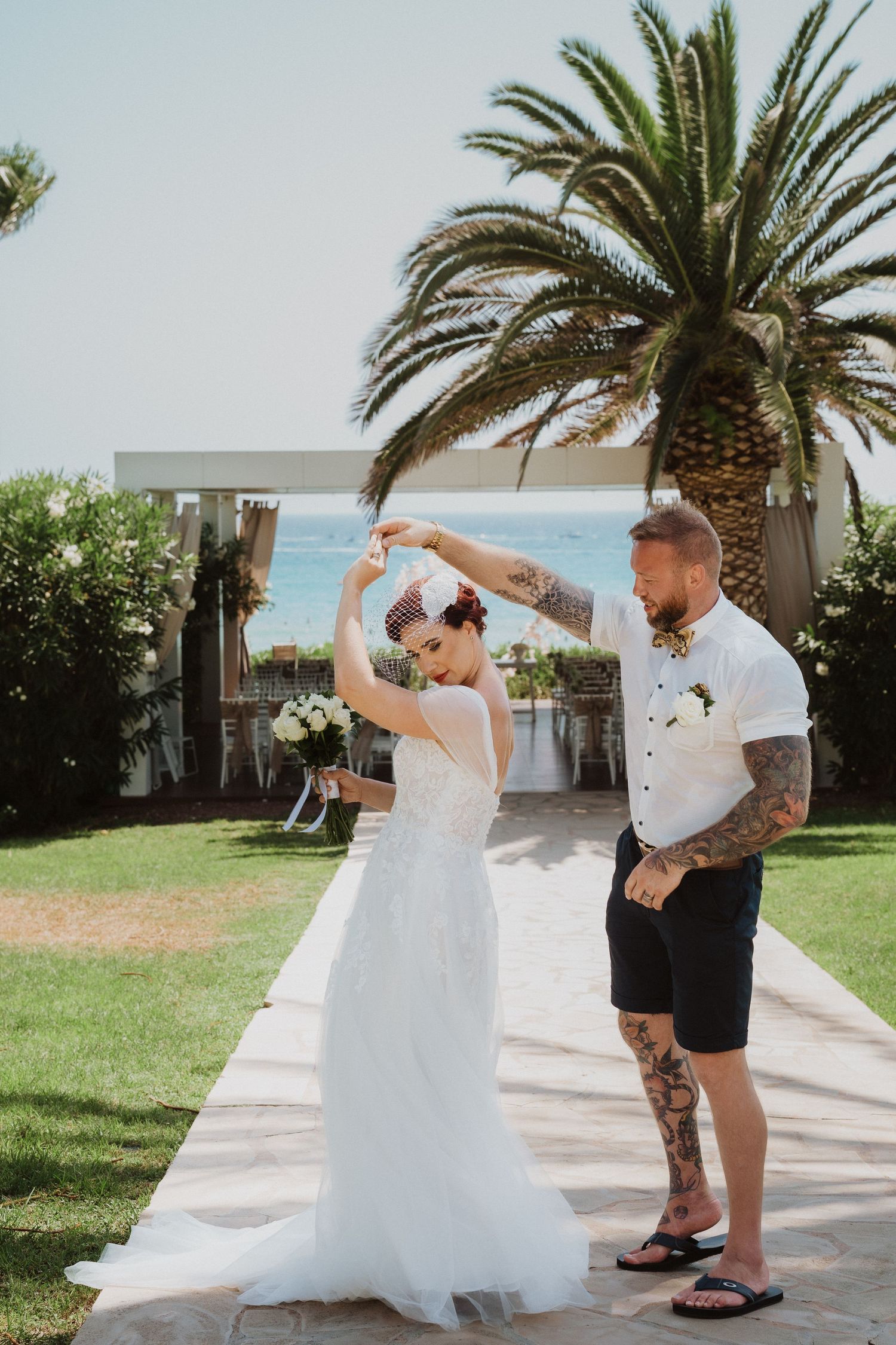 A wedding couple dances on a palm-lined pathway by the ocean on a sunny day.