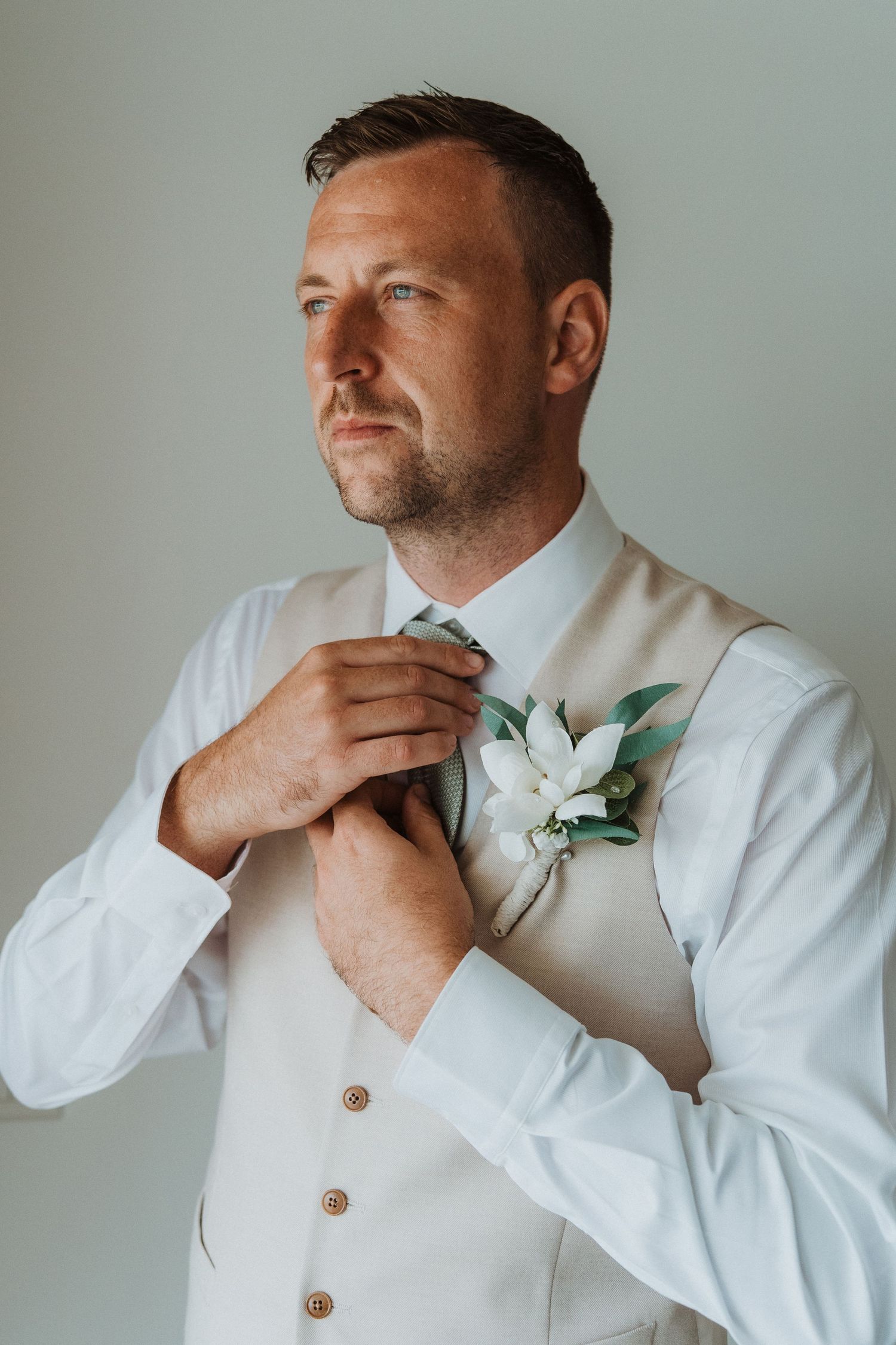 Groom in white vest and shirt adjusts tie while wearing delicate floral boutonniere against light background.