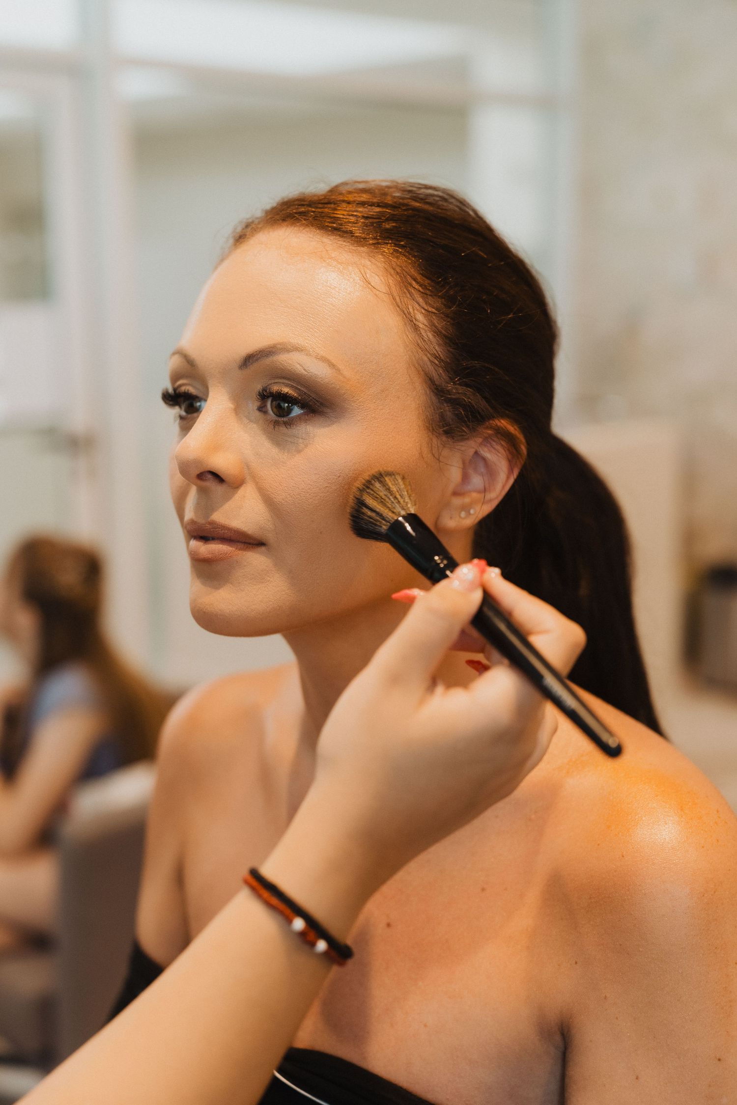 A makeup artist applies blush with a brush on a model during a beauty session in a bright studio setting.