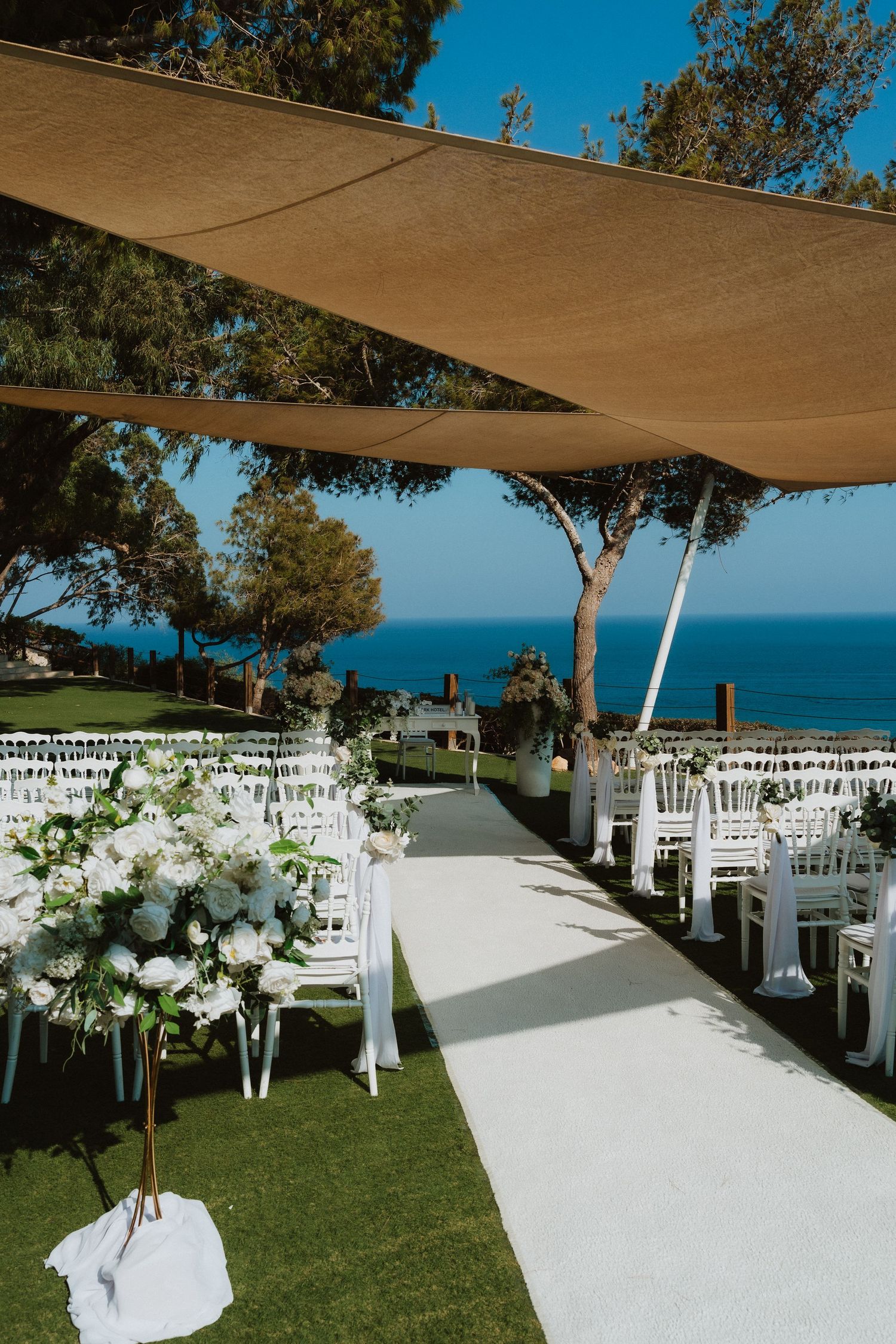 White wedding aisle with floral decorations leads to ocean view under beige shade canopy.
