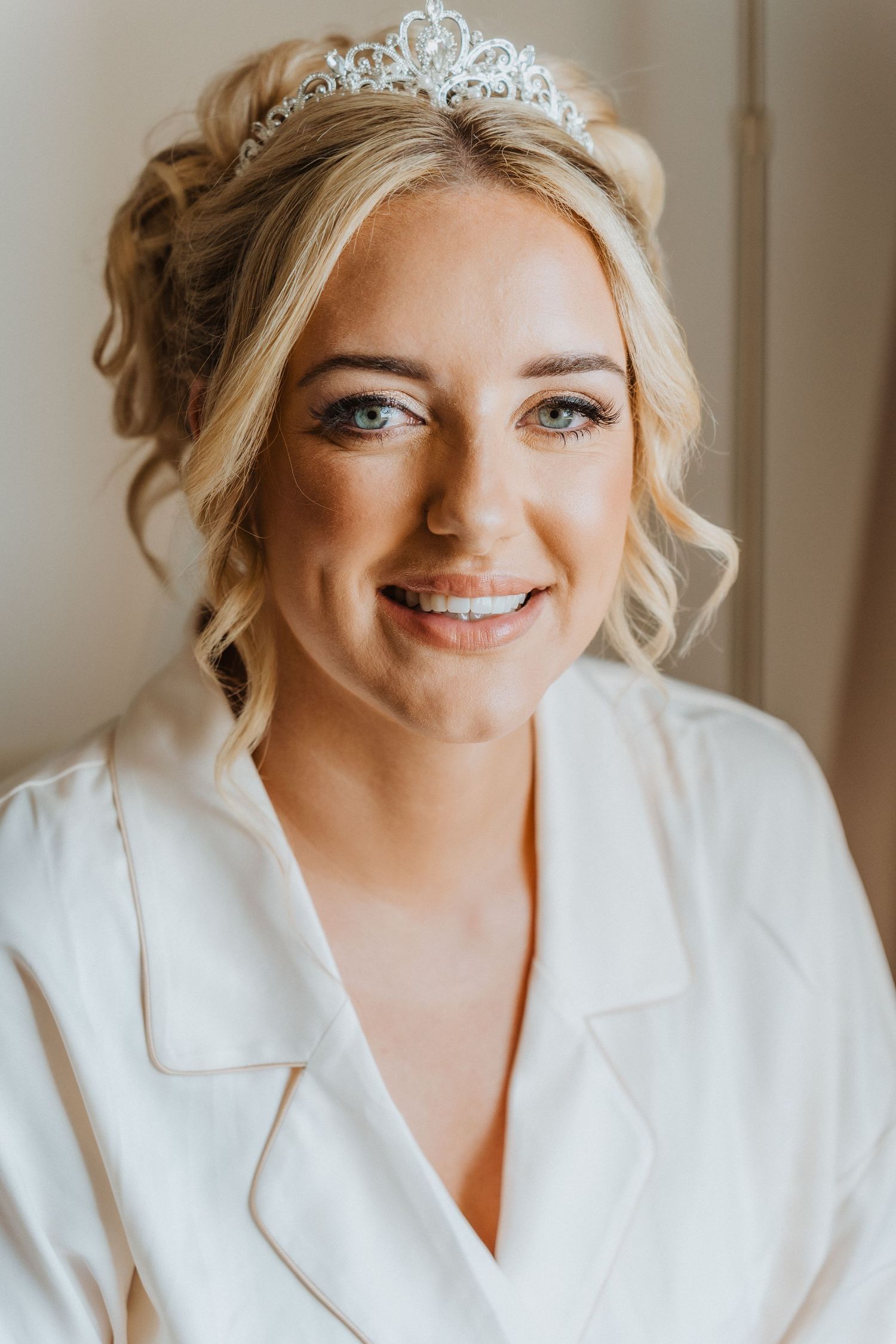 Bride wearing a delicate crystal tiara and white robe smiles warmly during wedding preparations.