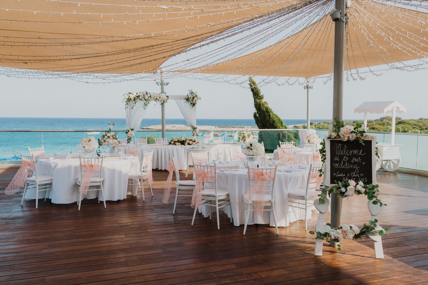 Elegant beachfront wedding reception setup with white chairs and coral accents under a draped canopy overlooking turquoise waters.