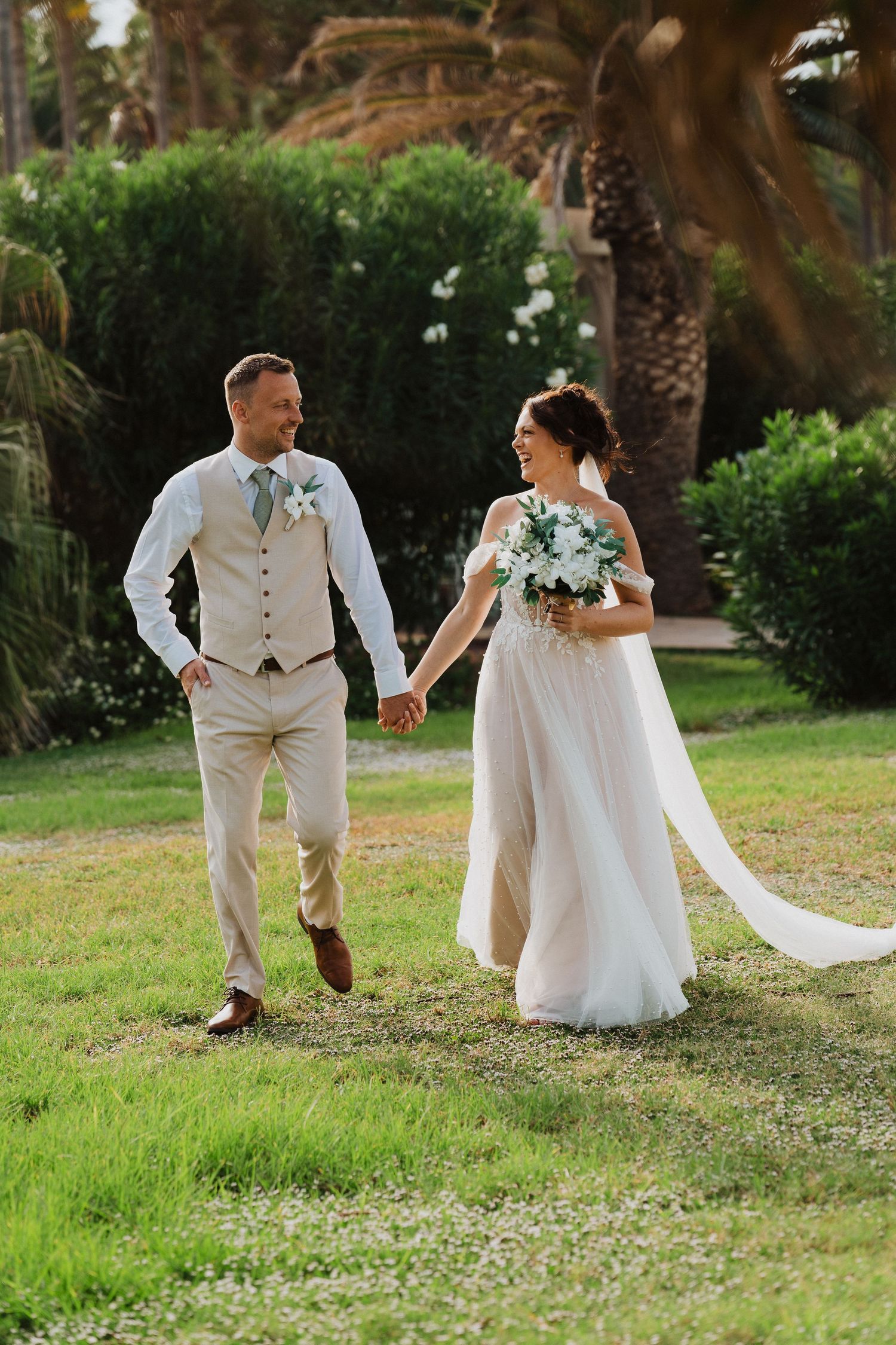 A couple holds hands while walking through a lush garden setting on their wedding day in light colored attire.