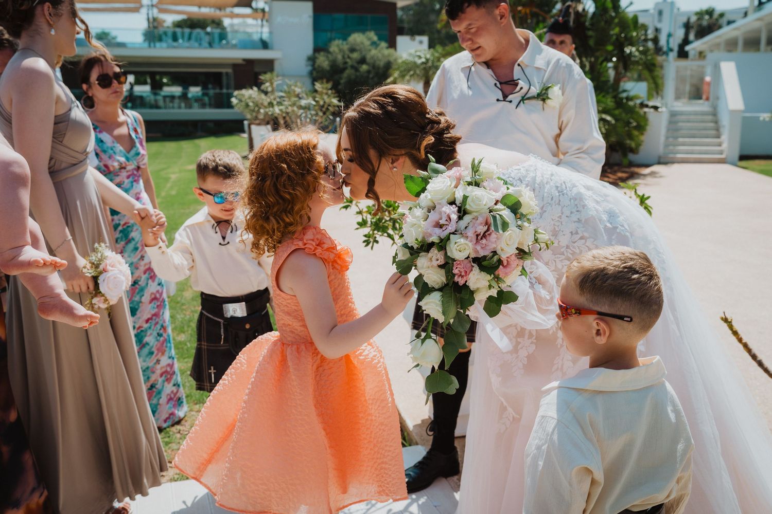 Little girl in peach dress admires bridal bouquet of white flowers at outdoor wedding.