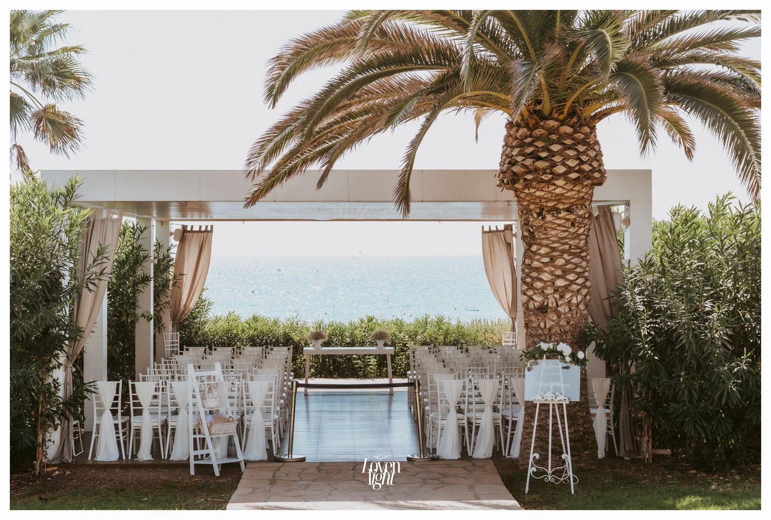 Outdoor wedding venue with white chairs and draped fabric arch framed by palm trees.