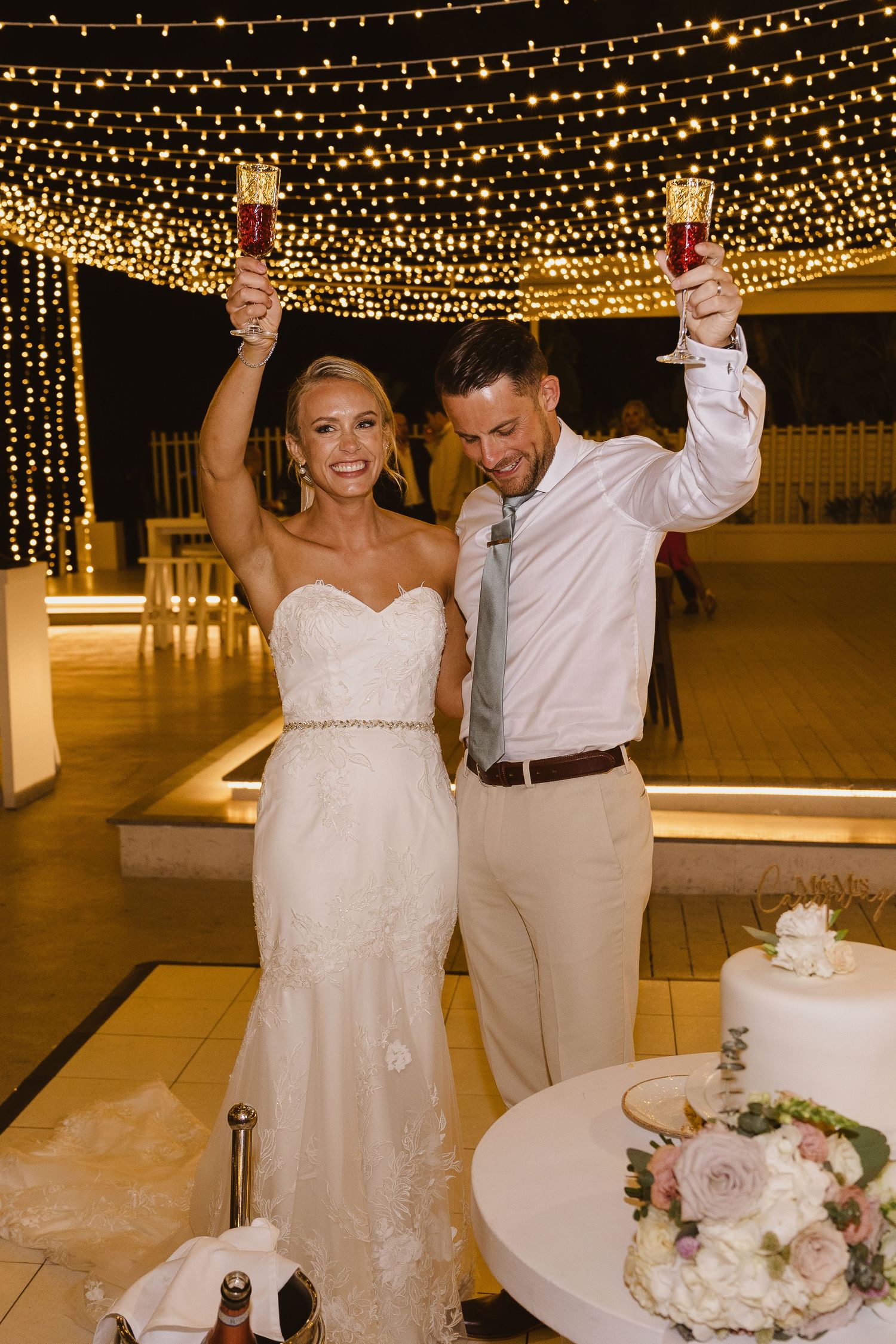 Wedding couple celebrating with raised glasses beneath a magical canopy of string lights.