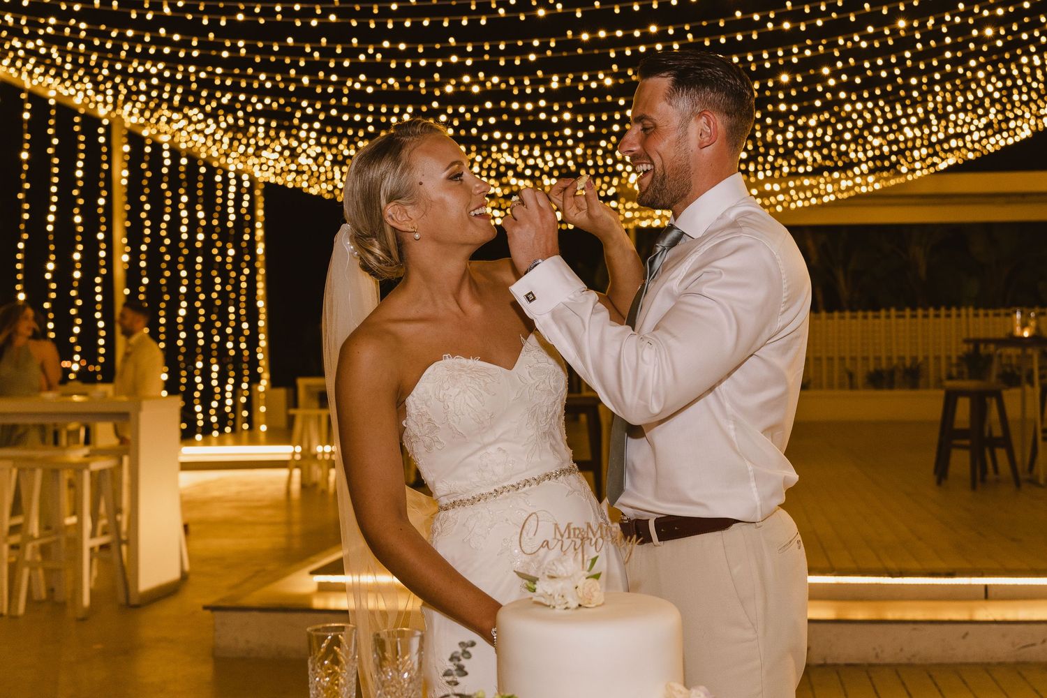 Wedding couple playfully feeds each other cake during reception under twinkling lights.