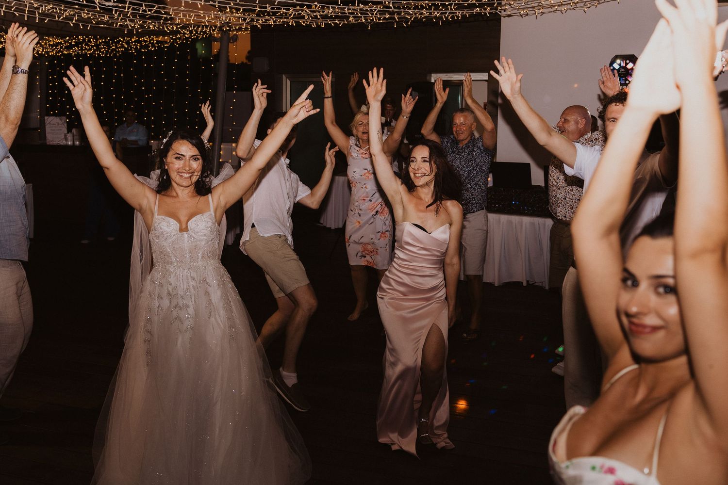 Wedding party dances energetically under canopy of twinkling lights during evening celebration.