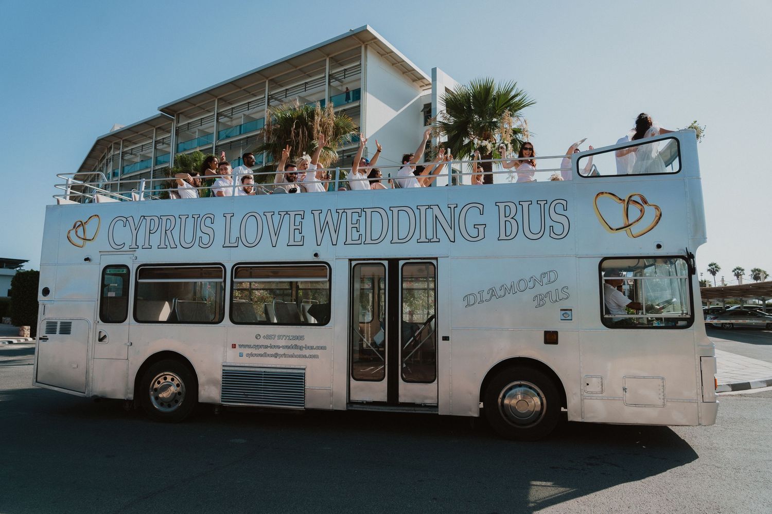 A light blue double decker wedding bus parked outdoors with celebratory decorations and passengers on top.