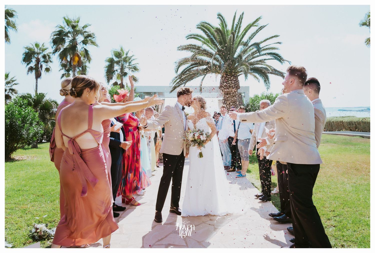 Guests throw confetti at an outdoor wedding celebration with tropical palm trees in the background.
