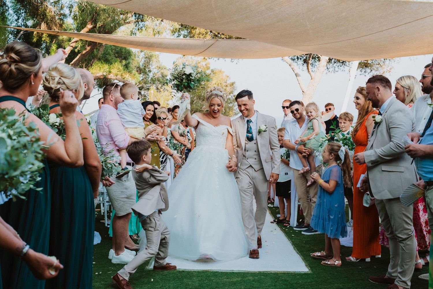 Wedding celebration under a canopy with guests cheering as couple walks down outdoor aisle.