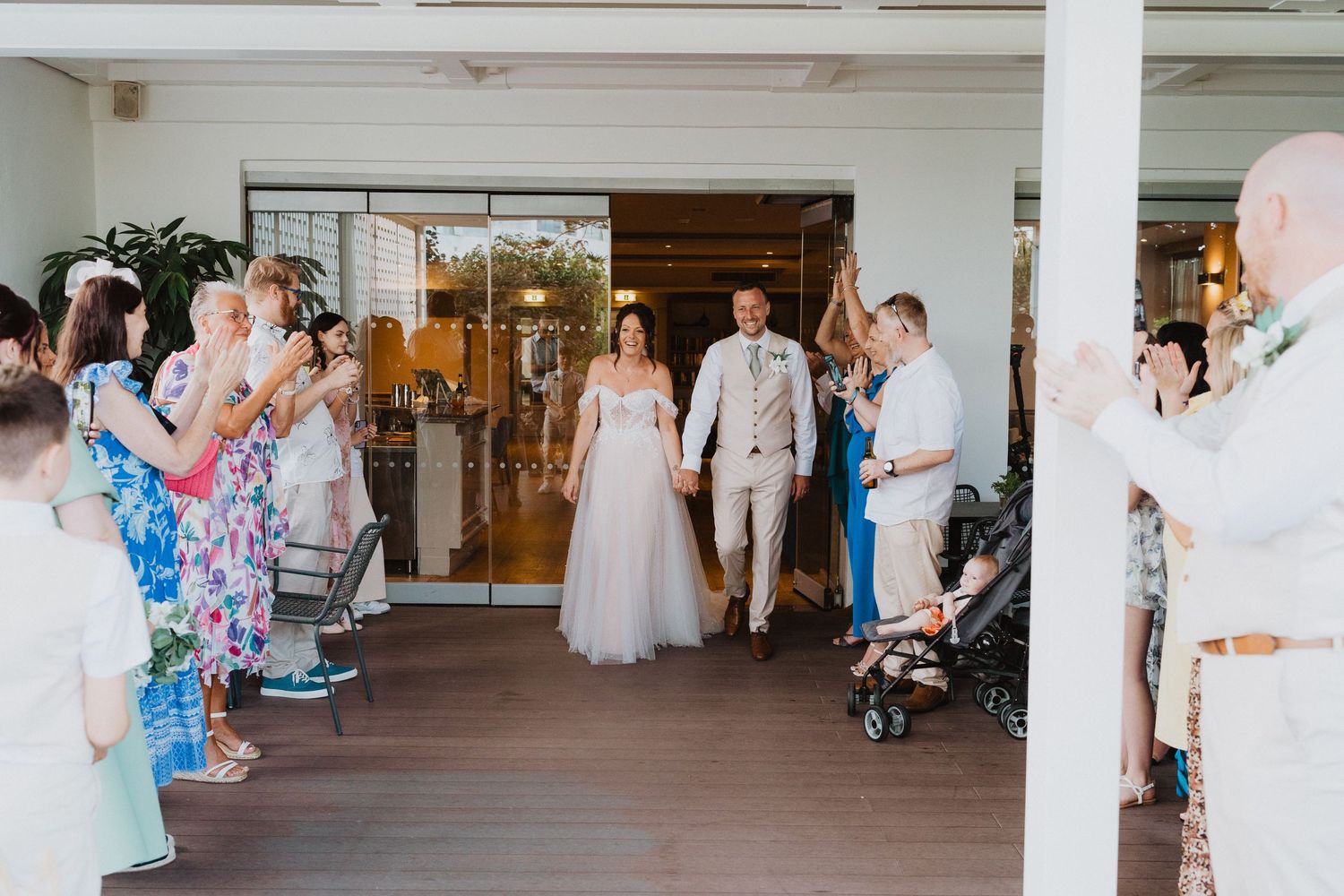 Wedding guests stand along a covered porch creating a walkway for the newlyweds exiting through glass doors.