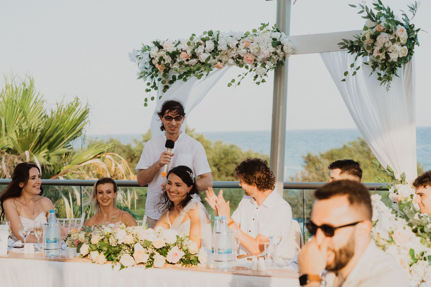 Wedding party seated at decorated head table during outdoor reception by the sea.