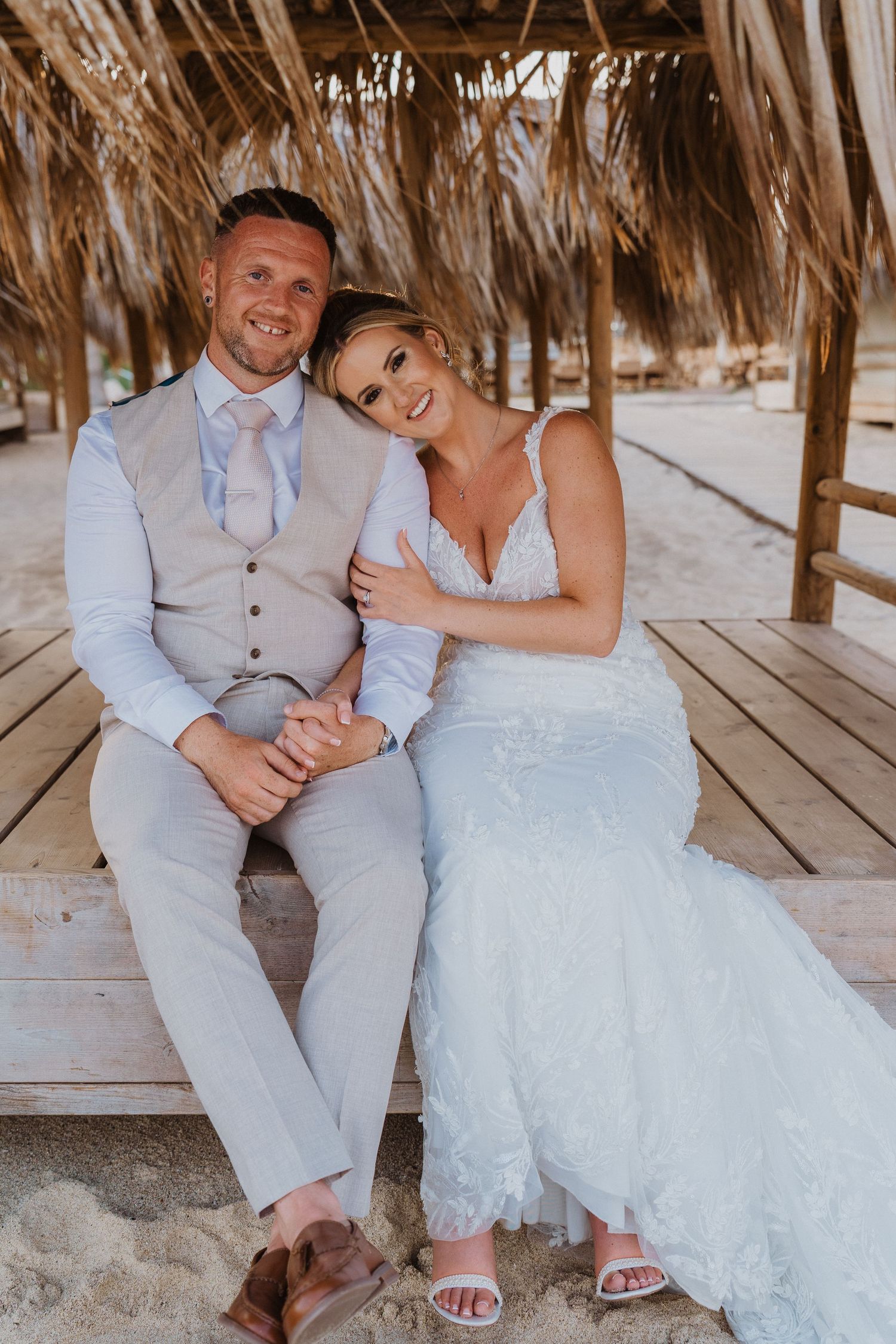 A couple in wedding attire sits together on a wooden deck at a tropical beach location.