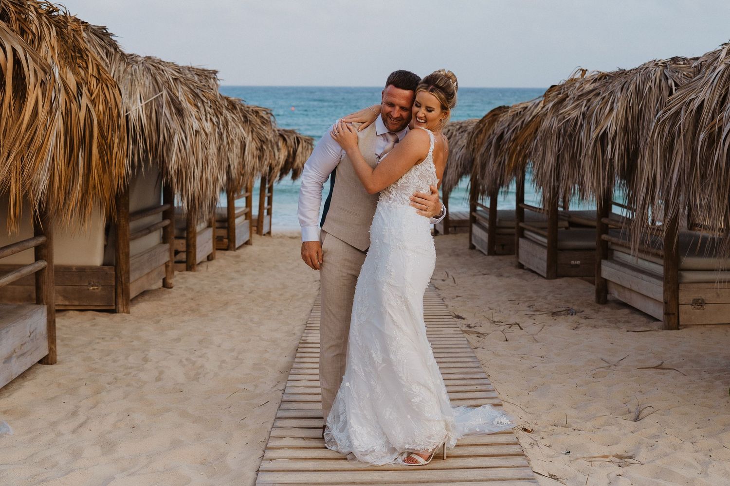 A romantic beach wedding scene with tiki umbrellas lining a sandy boardwalk path by the ocean.