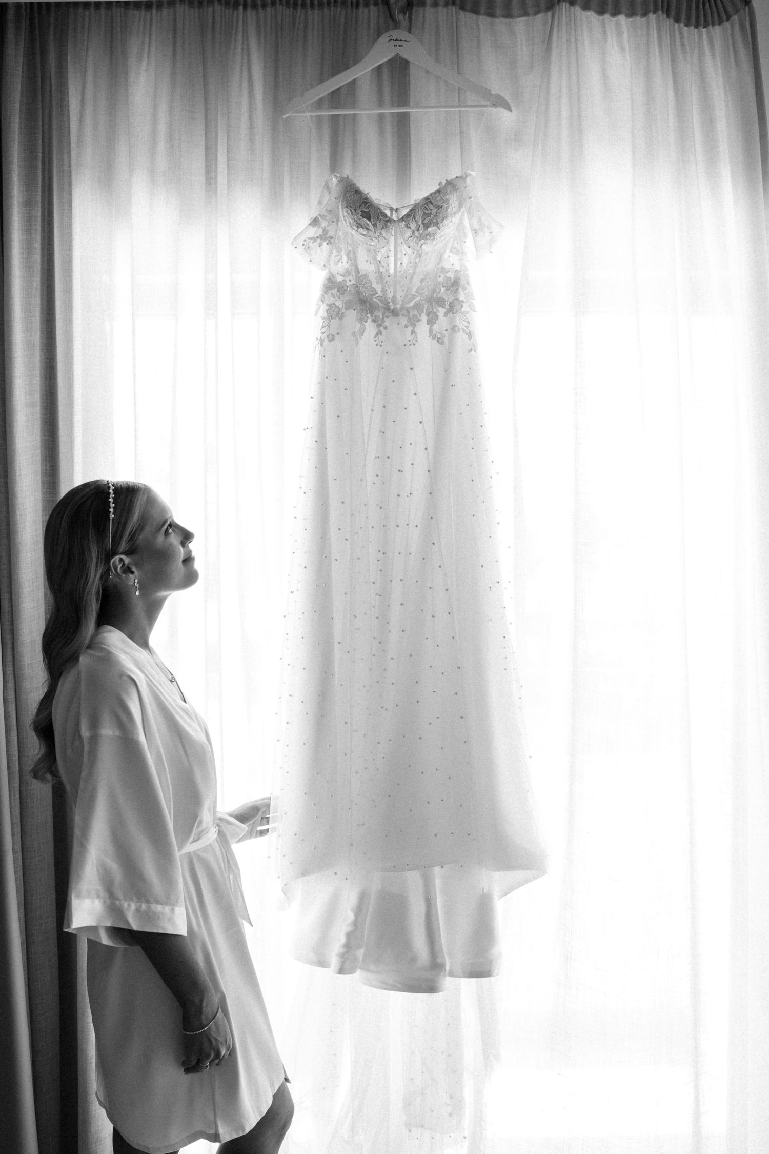Black and white photo of a wedding dress hanging by a window with silhouette in foreground.