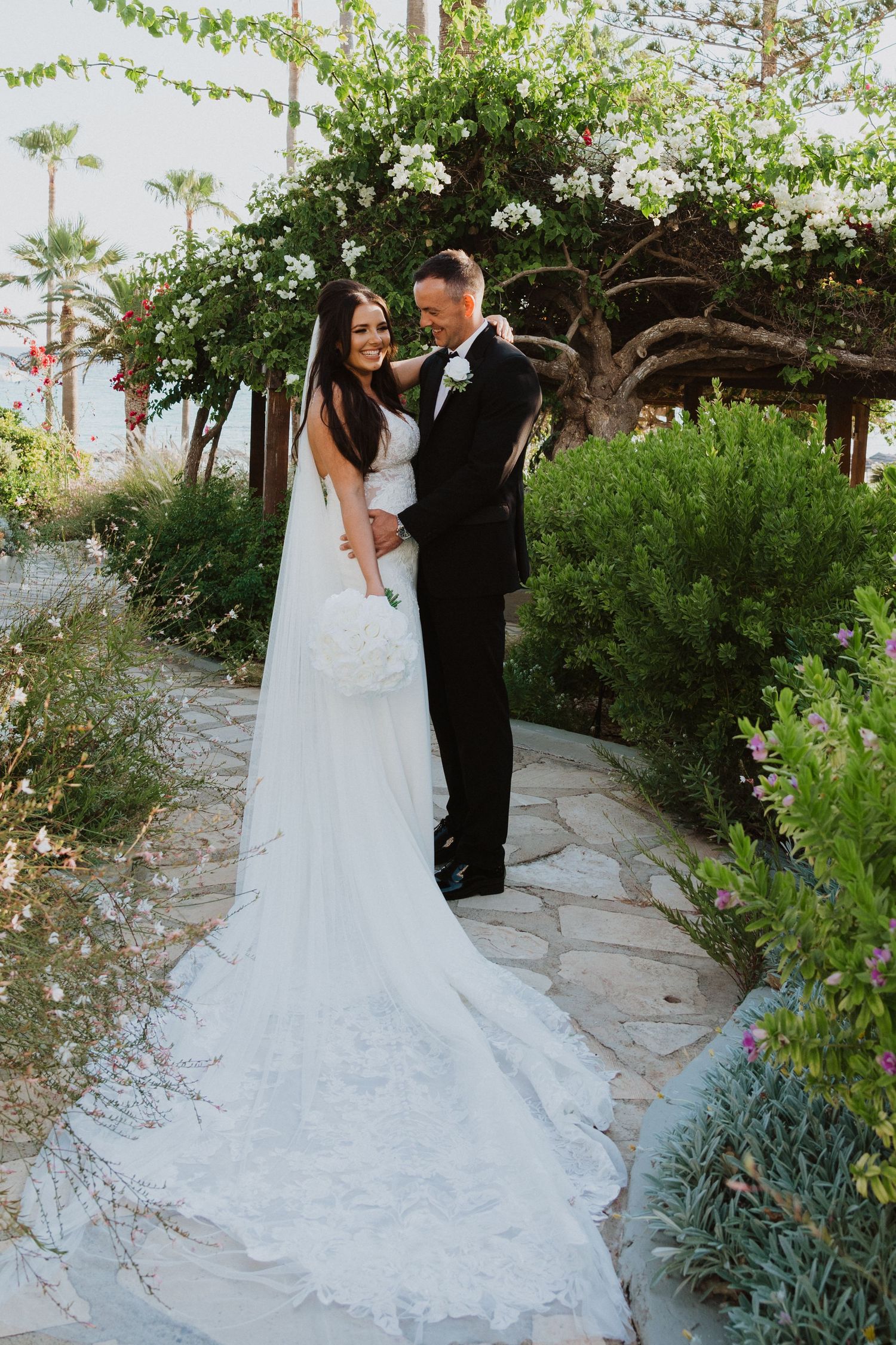 A romantic wedding scene unfolds on a winding garden path with palm trees and flowers.