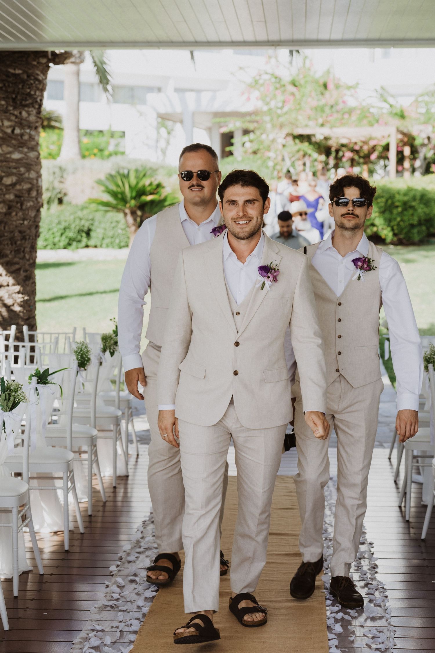 Groomsmen in light beige suits walk down a covered outdoor walkway decorated for a wedding.