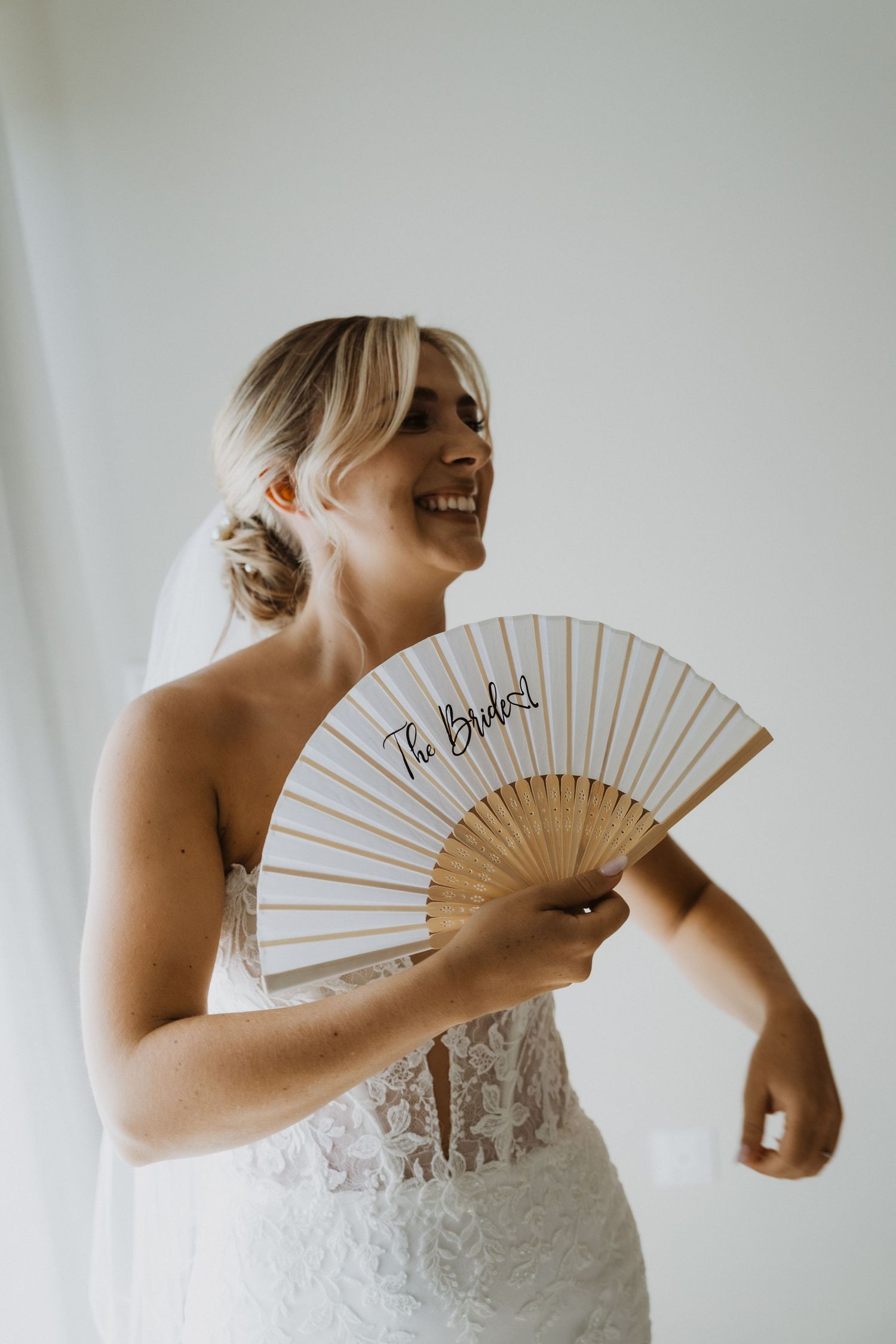 A bride in a white lace dress holds a decorative hand fan while smiling.