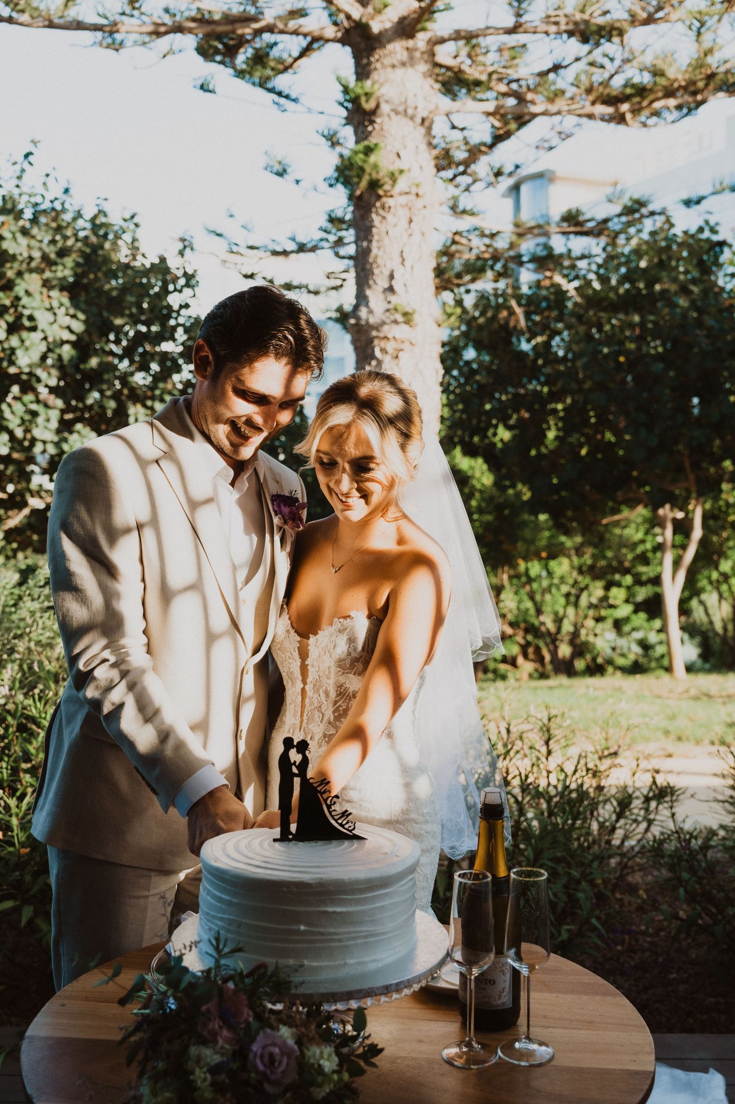 Newlyweds share a moment cutting their wedding cake outdoors under sunlit trees.