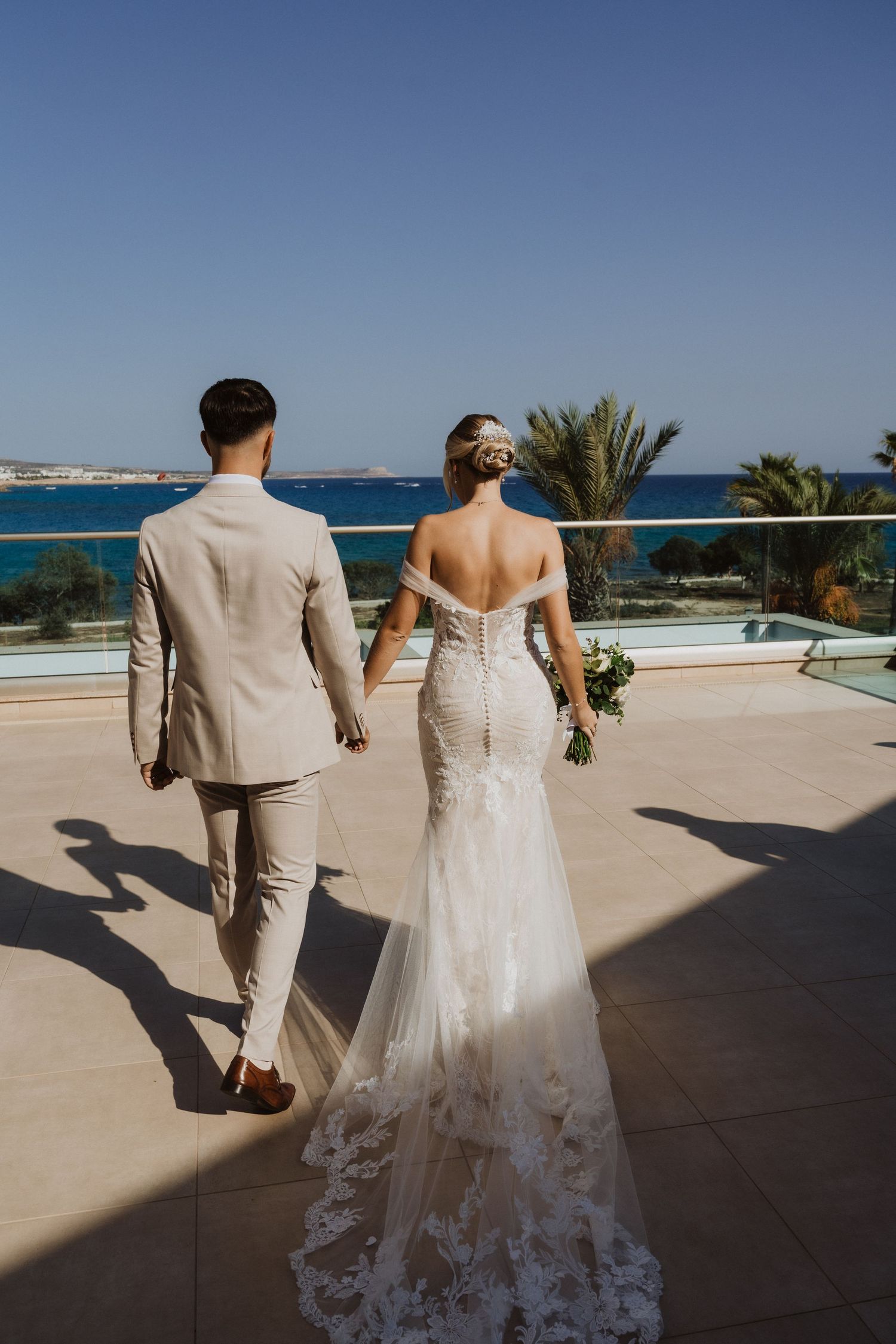 Newlyweds walking hand in hand on a sunny terrace with ocean views.