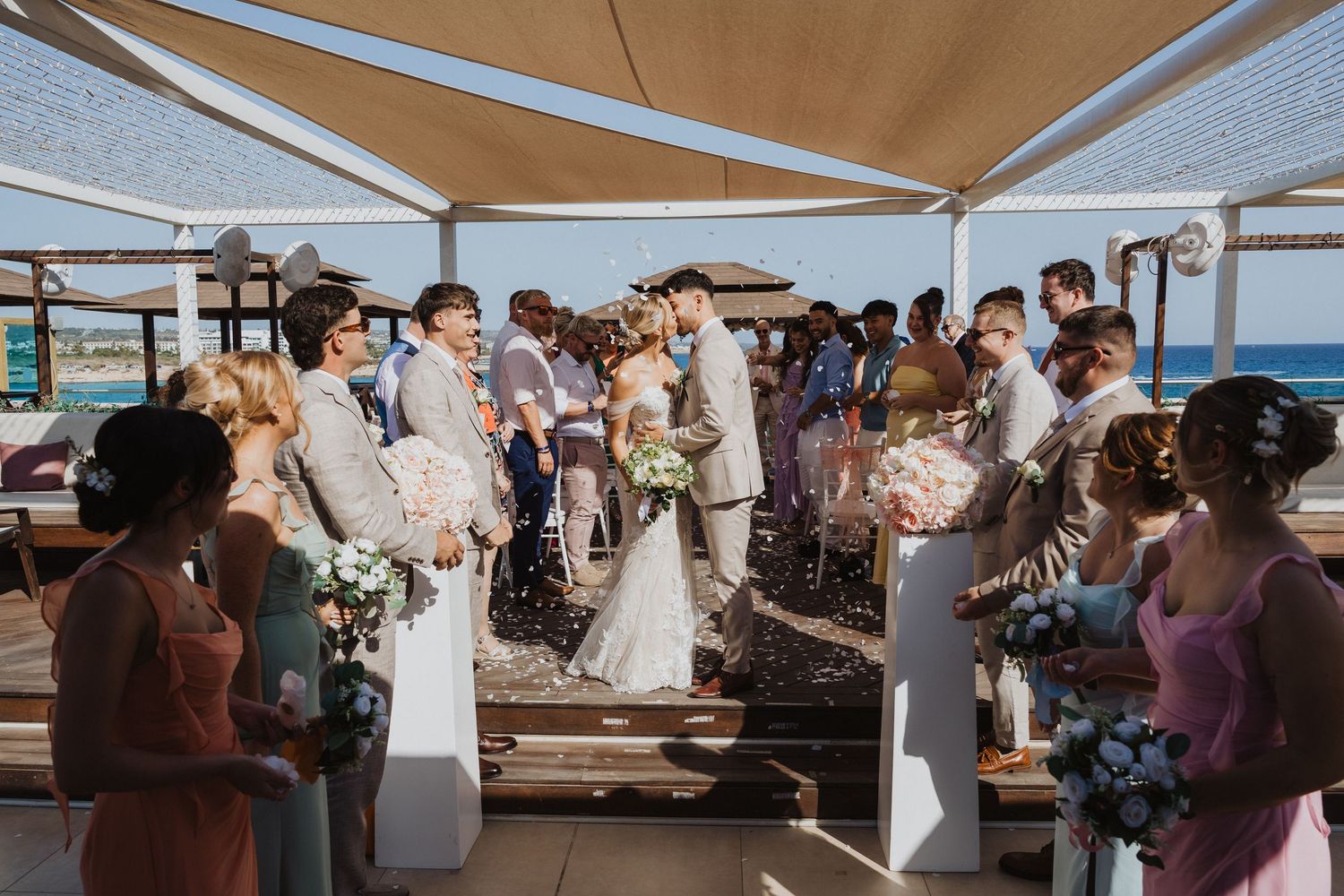 Wedding ceremony taking place under beige canopies at an outdoor waterfront venue with guests gathered around.