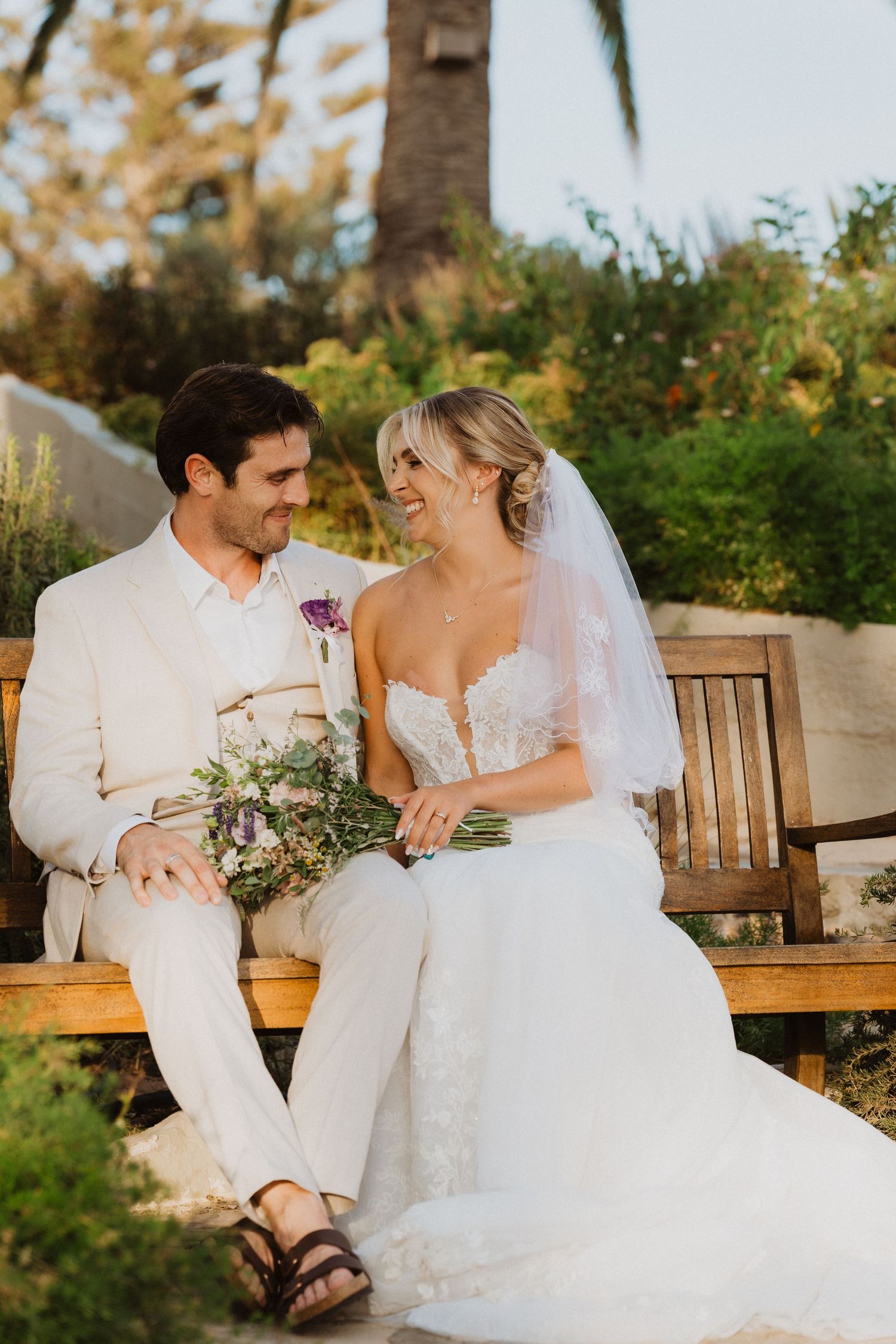 Bride and groom share candid moments while sitting on wooden bench in tropical garden setting.