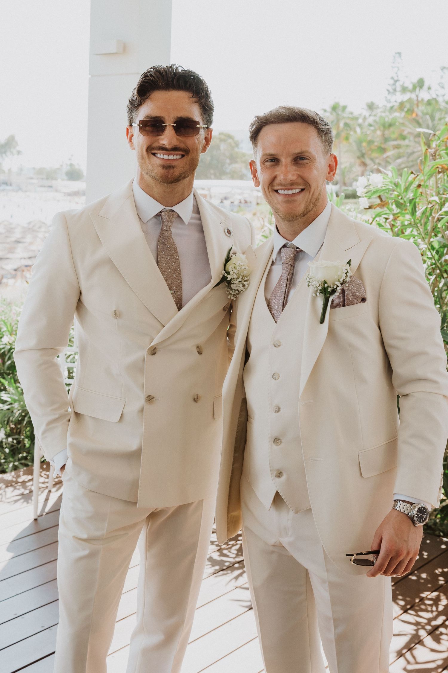 Two people in cream-colored wedding suits with boutonnieres smile together on a sunny outdoor porch.