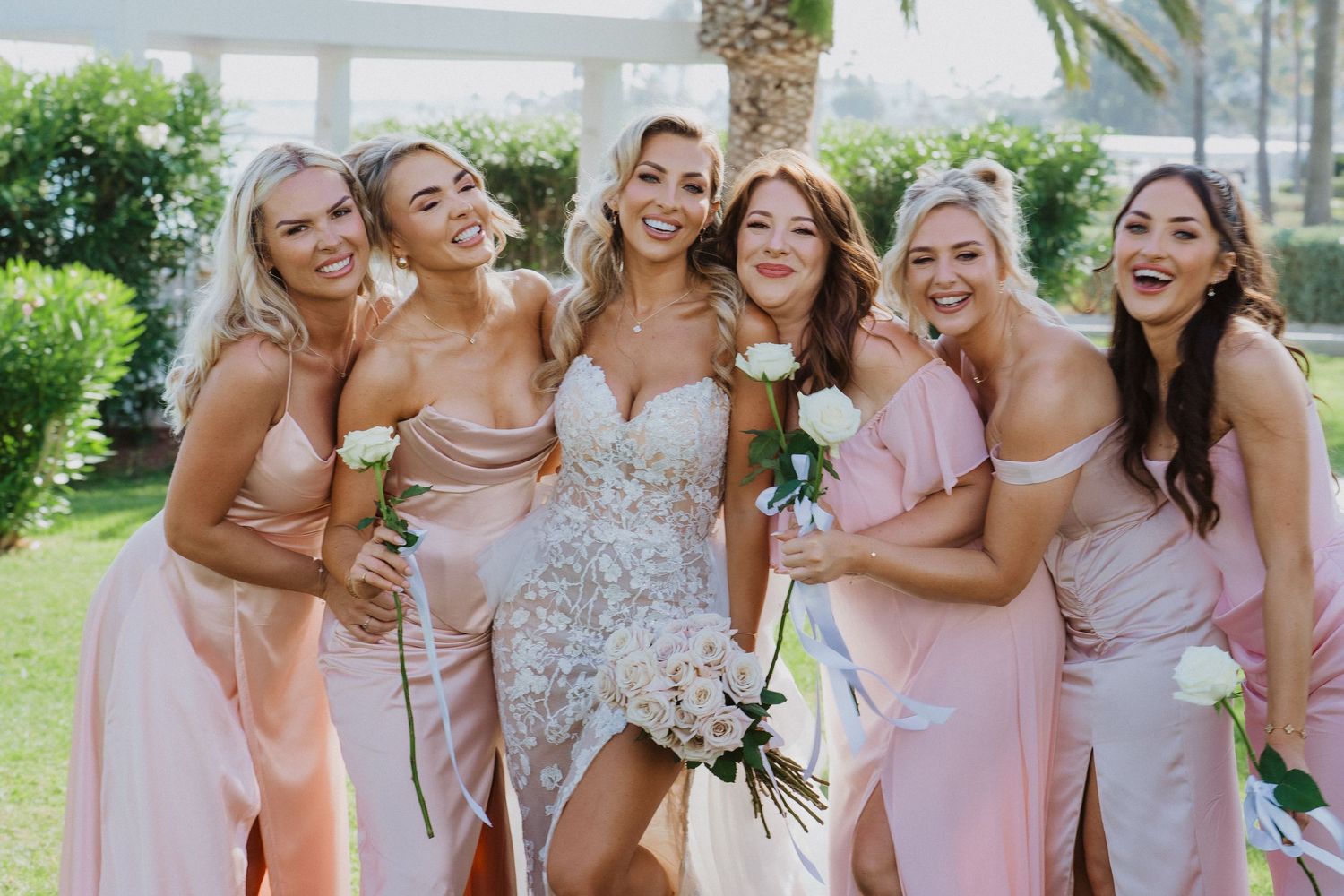 Wedding party in blush and pink gowns pose together while sharing joyful laughter at a garden celebration.