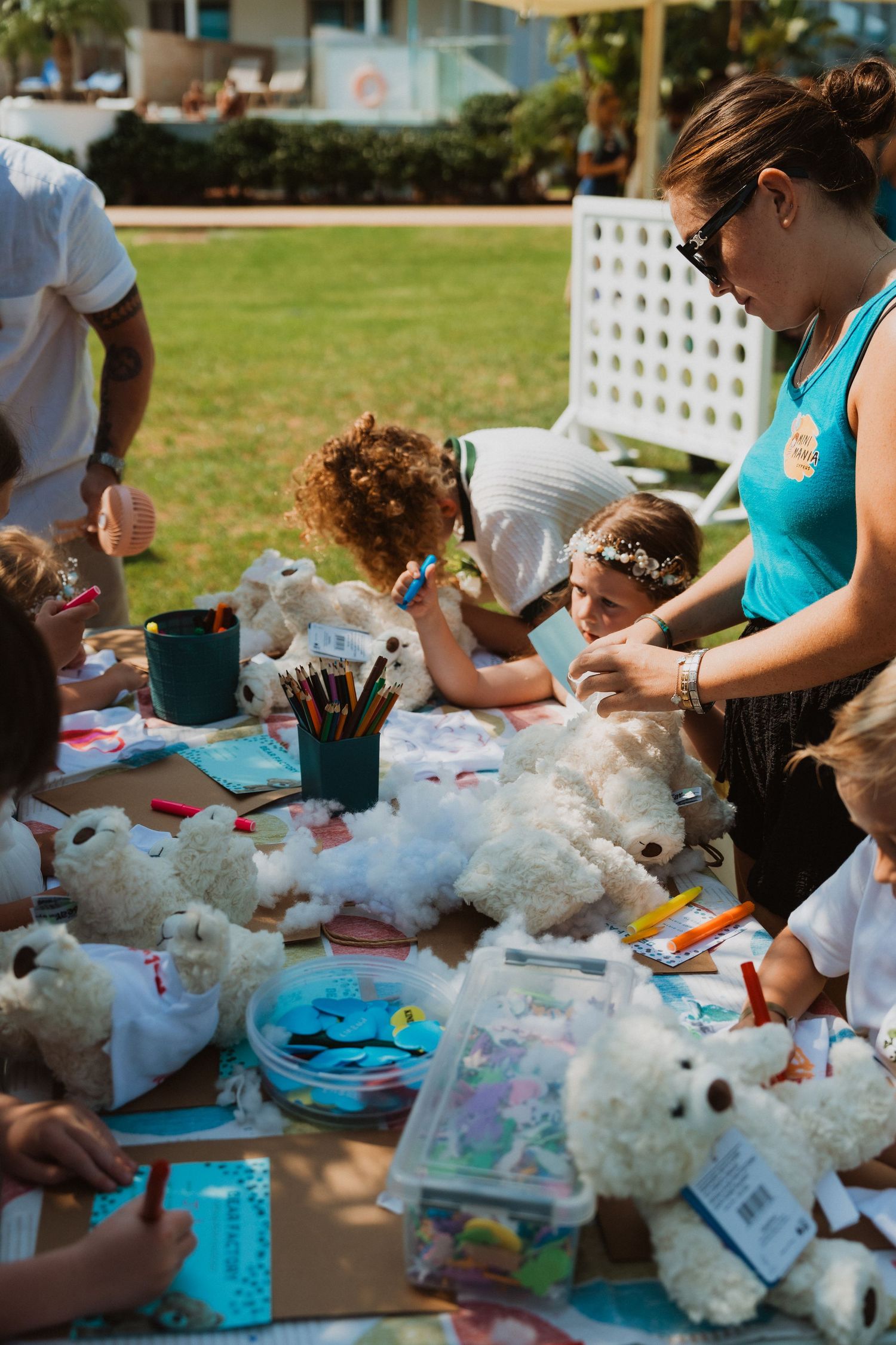 Children and adults craft stuffed teddy bears at an outdoor activity table during a sunny daytime event.