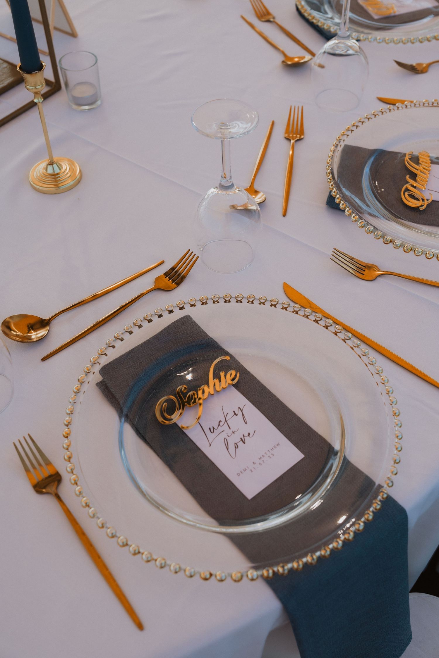 Elegant wedding table setting with gold cutlery, beaded charger plates, blue napkins, and personalized name cards.