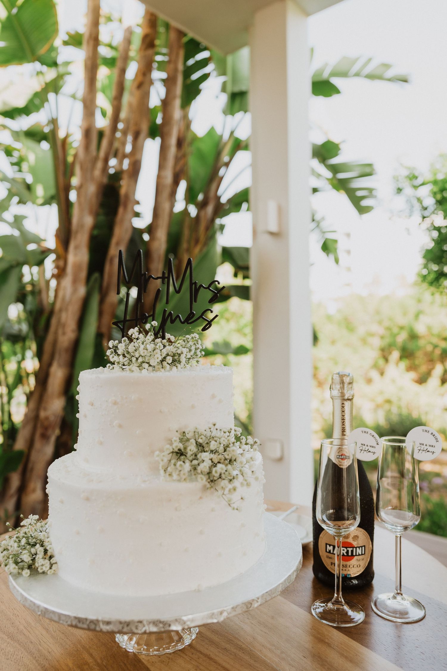 White two-tier wedding cake decorated with baby's breath flowers sits beside champagne bottle and glasses on wooden table.