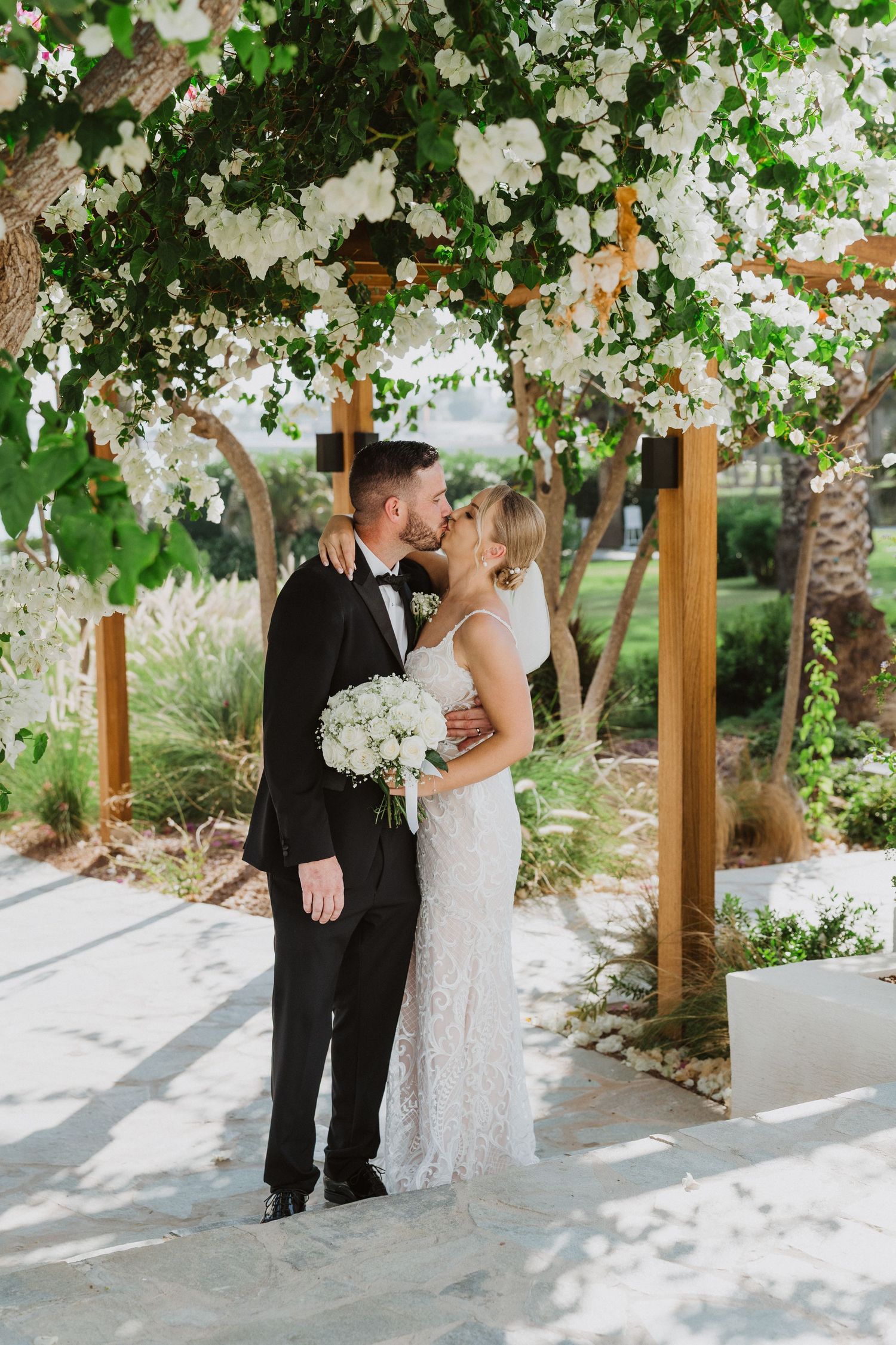 Wedding couple embracing beneath white flowering vines and wooden beams in a garden setting.