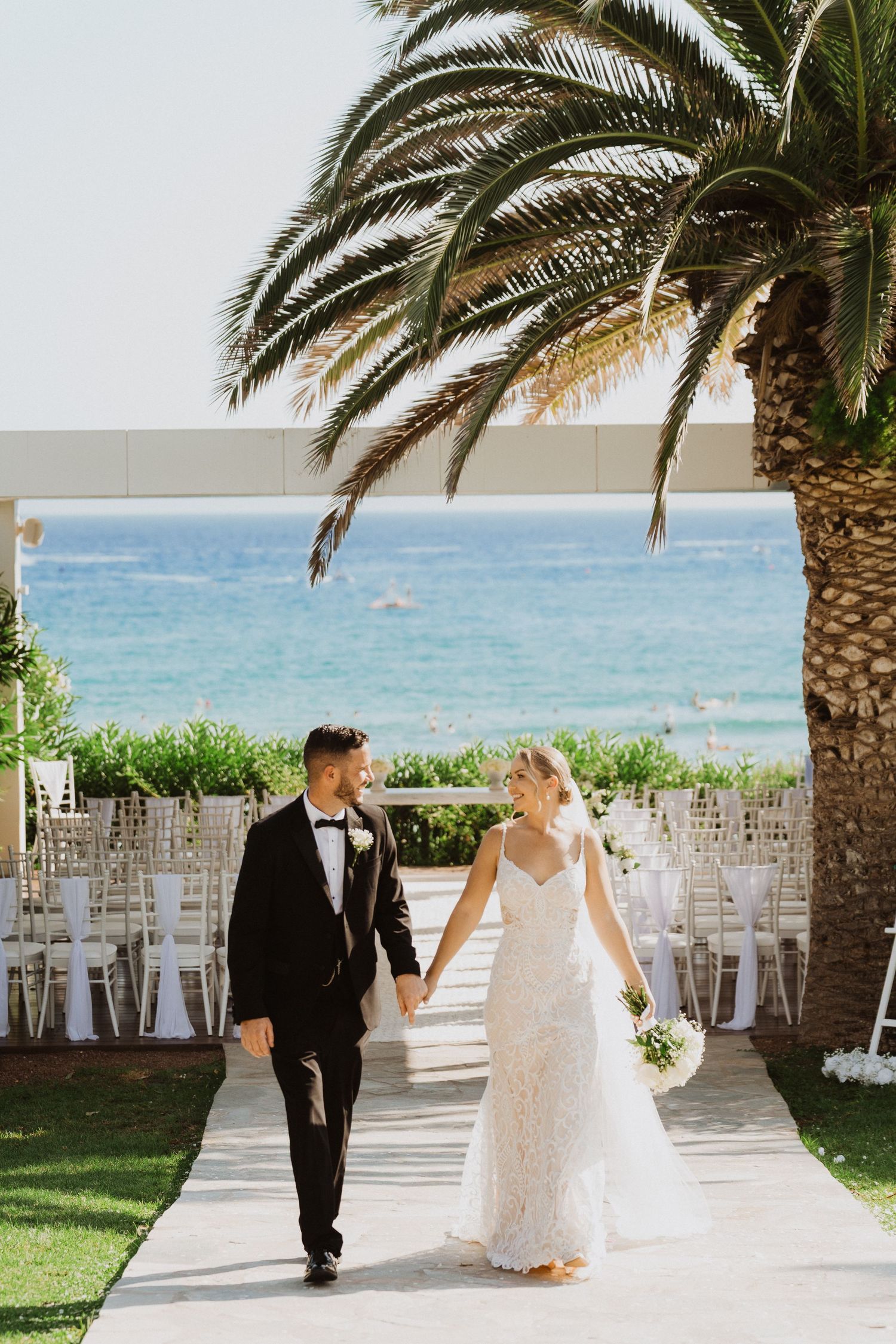 Bride and groom share romantic moments under palm trees at a beachside wedding venue with white chairs and ocean views.