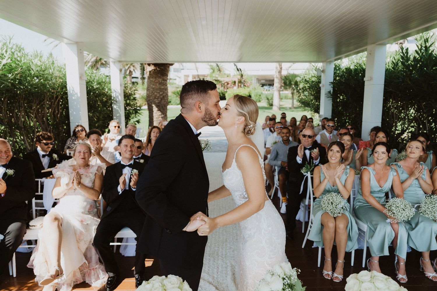 Wedding party members in light blue dresses and black tuxedos pose together at an outdoor celebration.